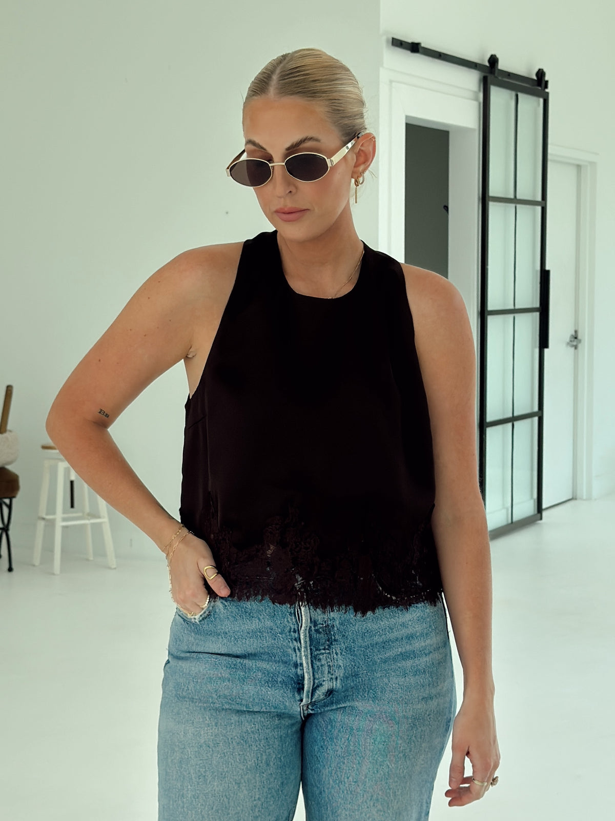 A woman modeling a sleeveless brown top with a textured, frayed hem and lace-like trim, paired with light-wash blue jeans in a bright, modern indoor setting.