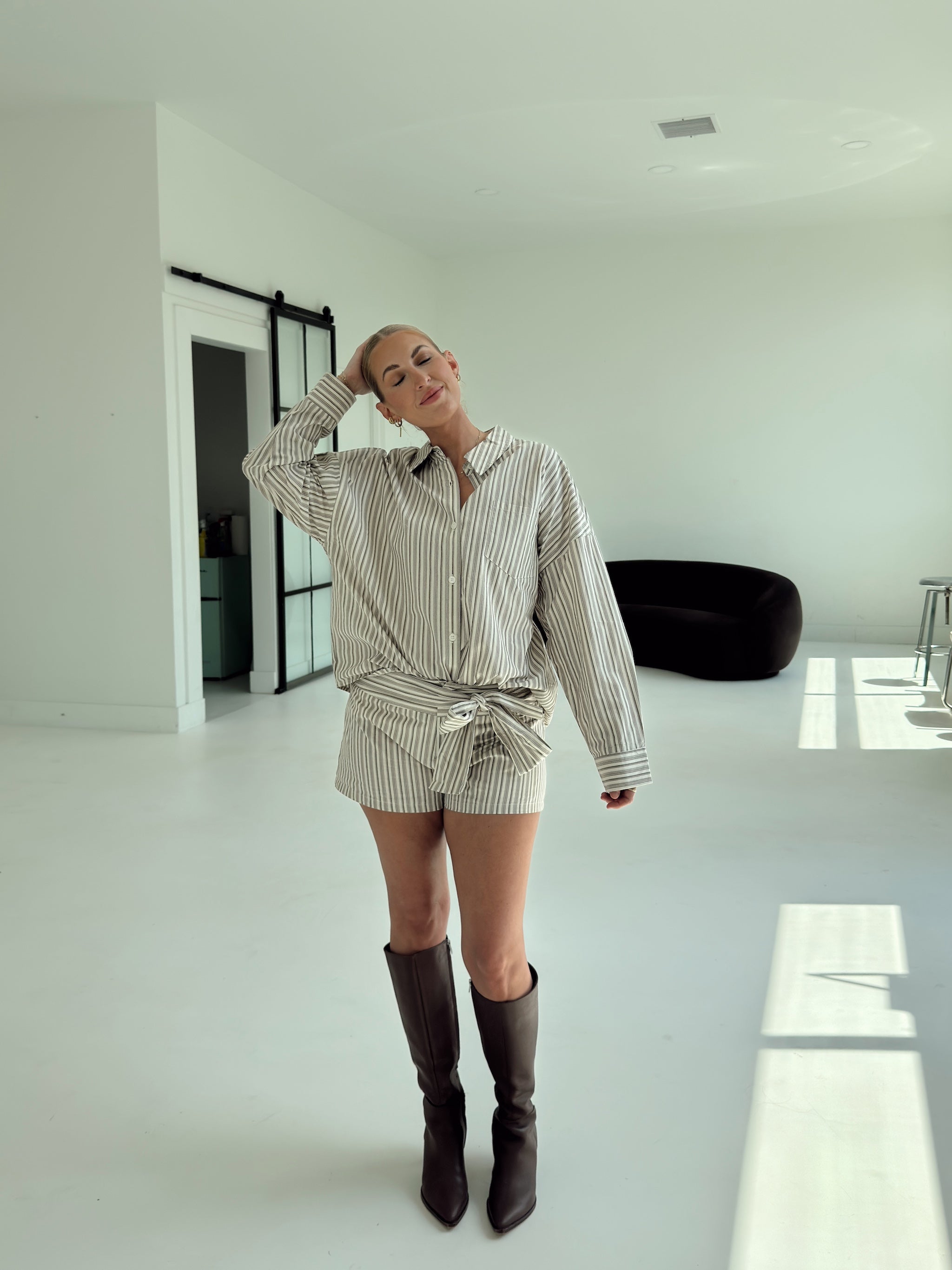 A woman models a matching two-piece short-sleeve set consisting of a button-down shirt and shorts in a beige and white vertical stripe pattern, tied at the waist. She is wearing dark brown knee-high boots in a bright, modern white room with a black curved lounge chair in the background.