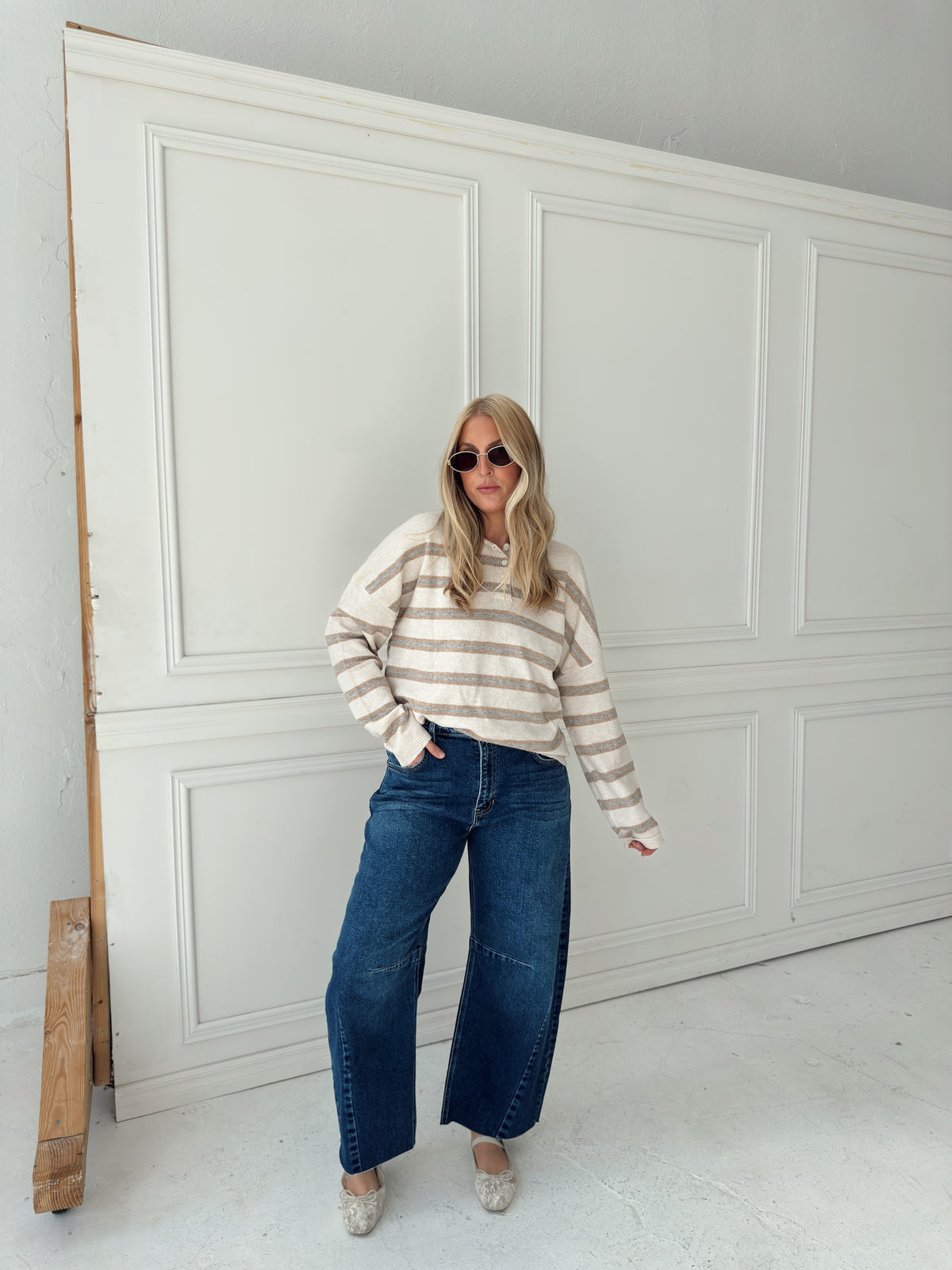 A woman stands in front of a white paneled wall wearing a beige, cream, and light blue horizontally striped long-sleeve knit top, wide-leg blue jeans, and light-colored flats.