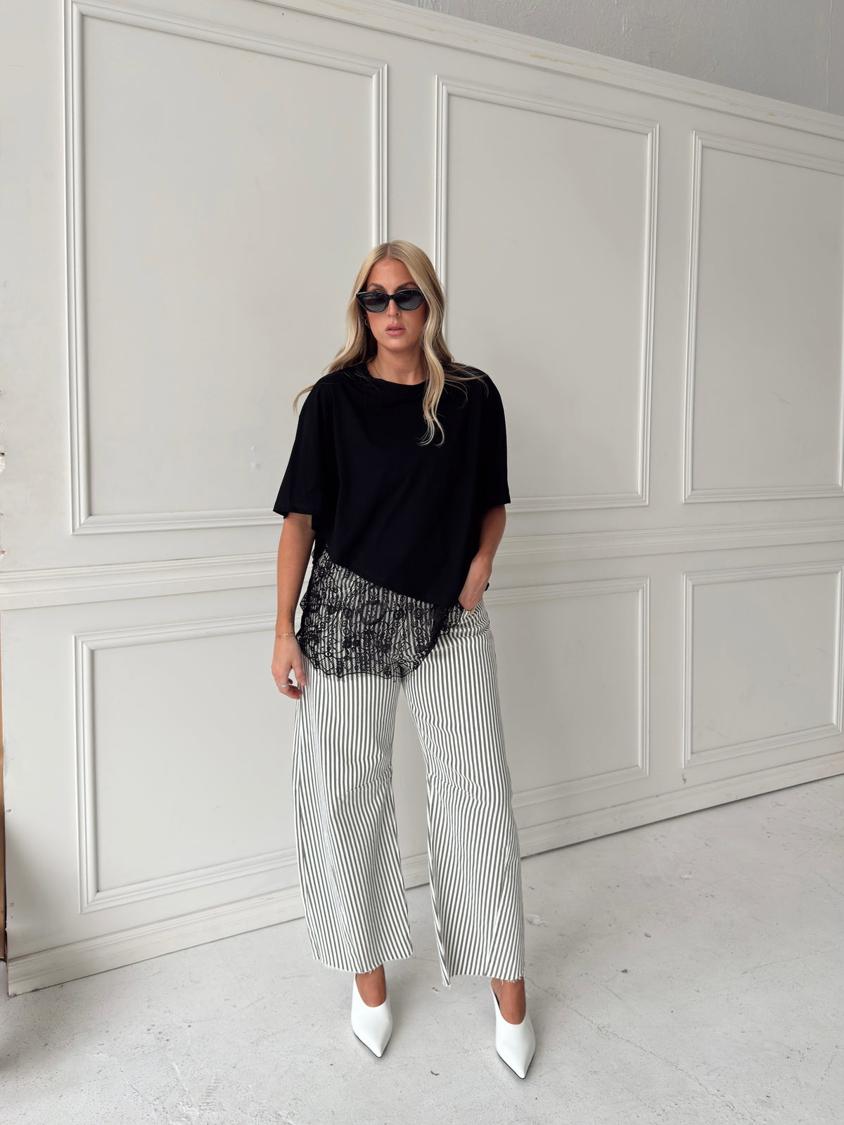 A woman modeling a pair of wide-leg barrel jeans with thin olive and white vertical stripes, paired with a black t-shirt, black sunglasses, and white pointed-toe heels, standing against a white paneled wall.