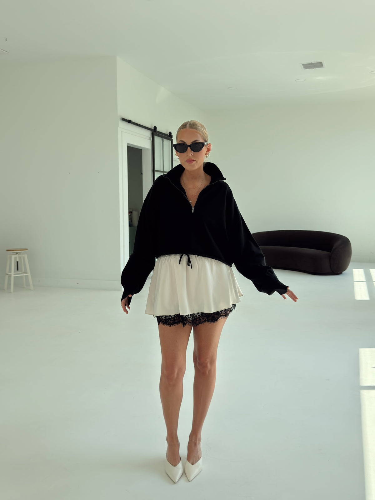A woman modeling a black zip-up hoodie and a white ruffled mini skirt with black lace trim, styled with white pointed-toe heels and black sunglasses in a bright, minimalist white room.