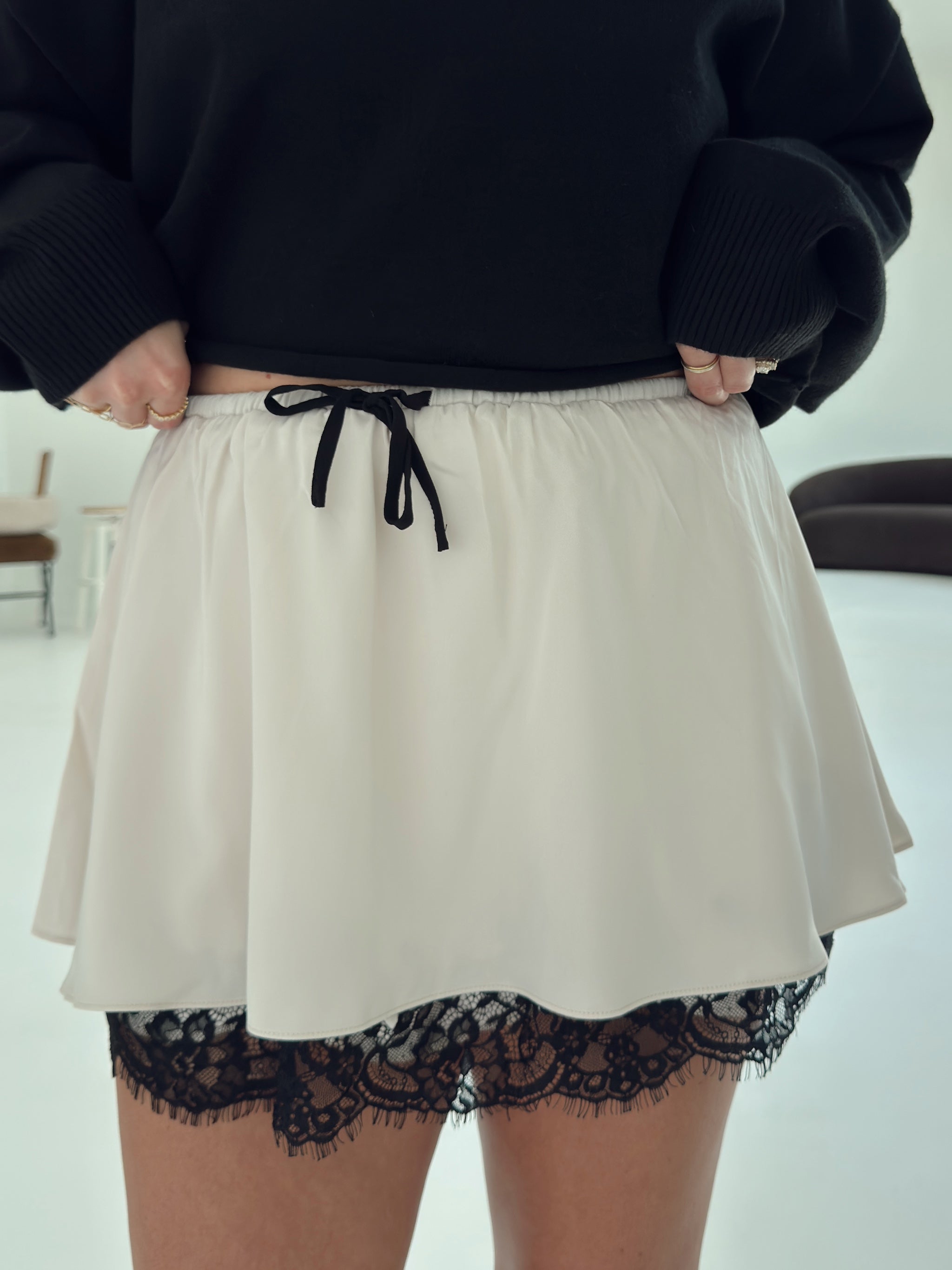 A person wearing a short cream skirt with a black drawstring and black lace trim at the hem, paired with a black sweater. The photo is cropped from the chest down in an indoor setting.