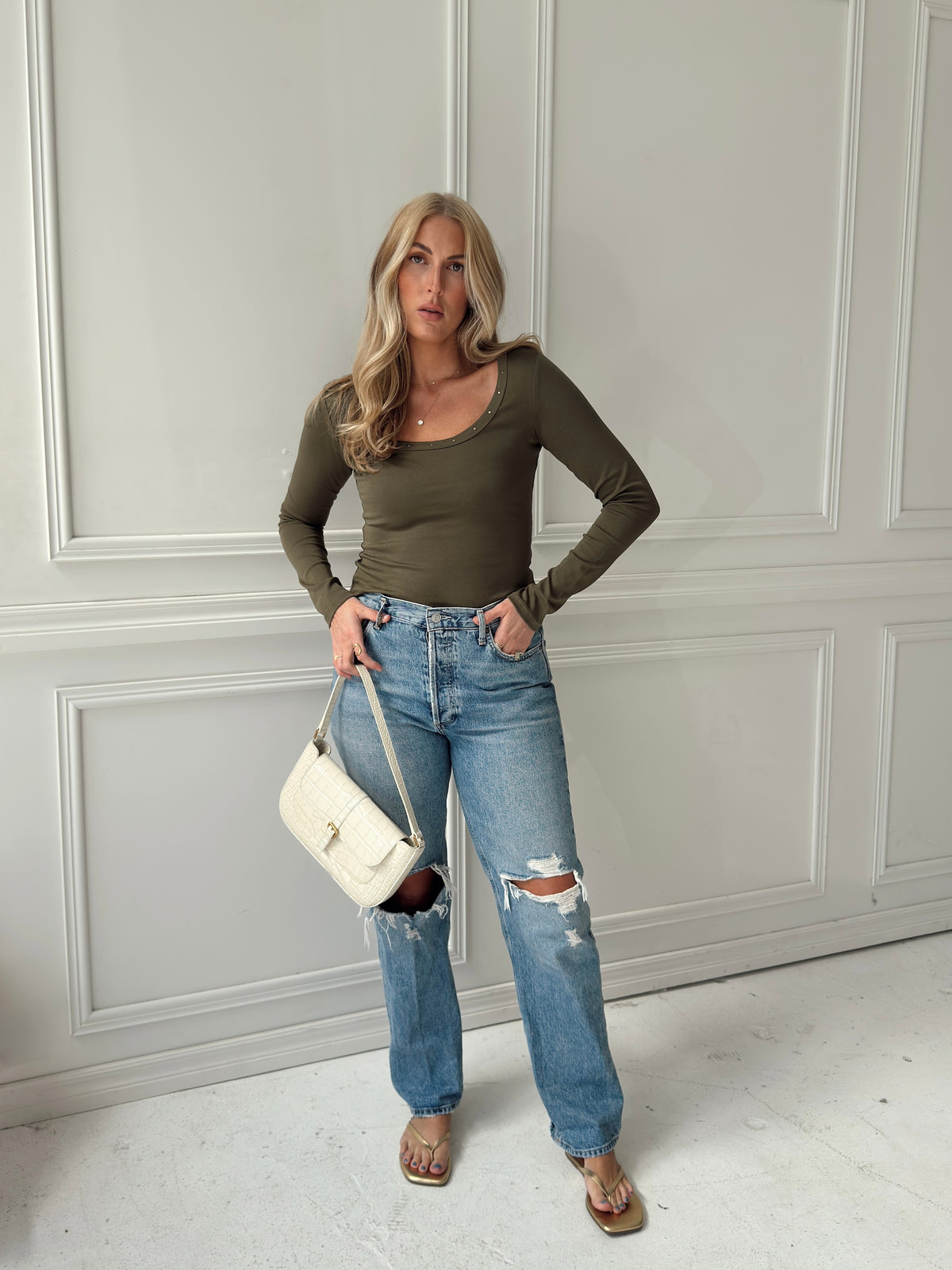A woman stands indoors against a white paneled wall wearing an olive green long-sleeve scoop-neck top, distressed light-wash blue jeans, and tan sandals, while holding a white shoulder bag.