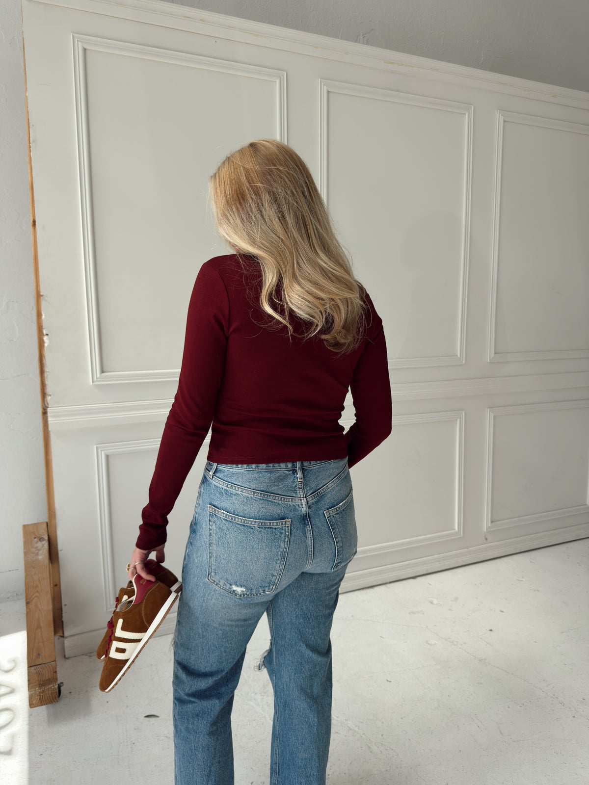 A woman with blonde hair is seen from behind, wearing a burgundy long-sleeve top and blue jeans, holding brown sneakers in a room with white paneled walls.
