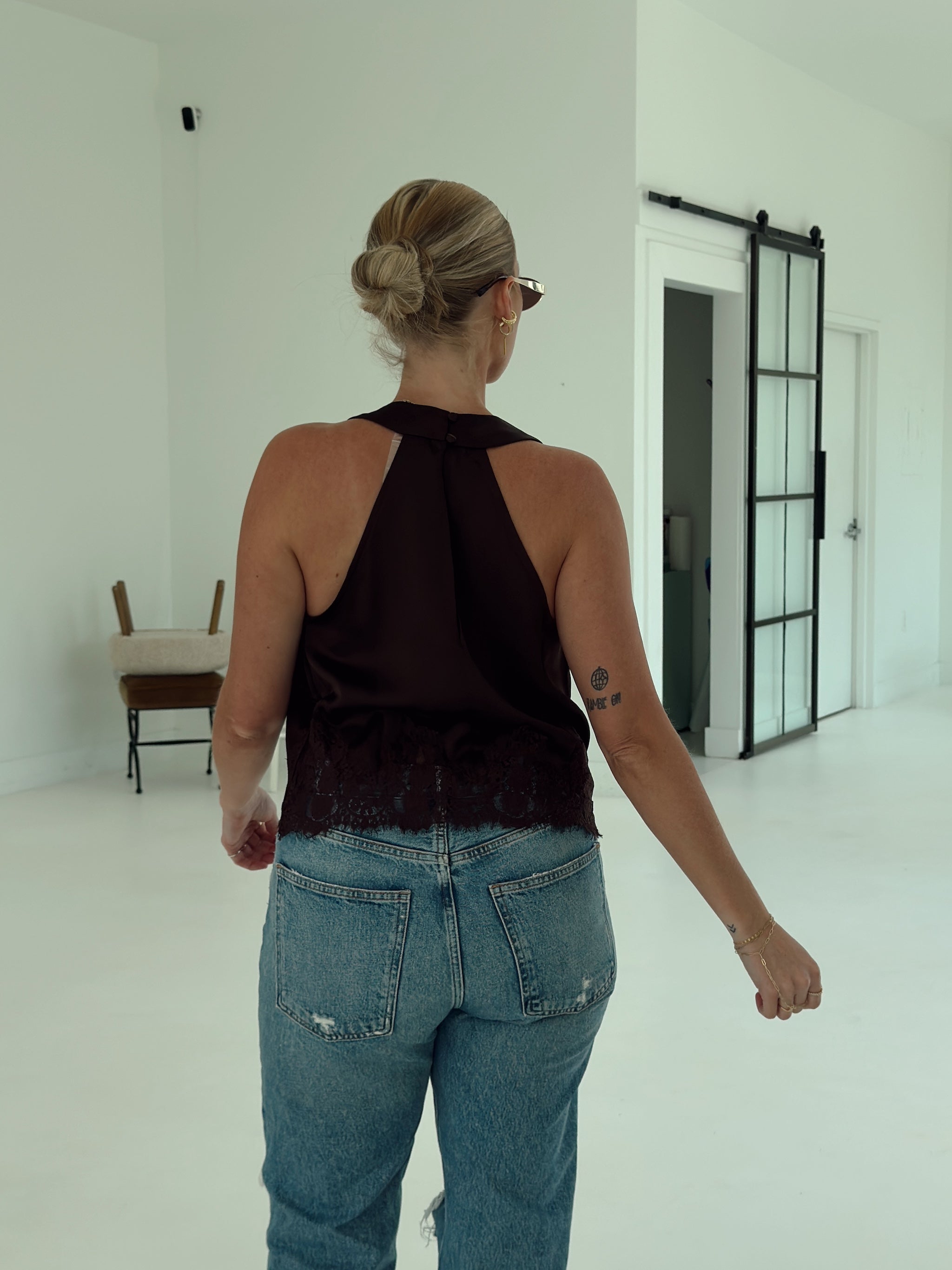 A woman viewed from behind wearing a dark brown halter top with a ruffled lace hem and blue jeans, standing in a bright white room with a sliding glass door in the background.