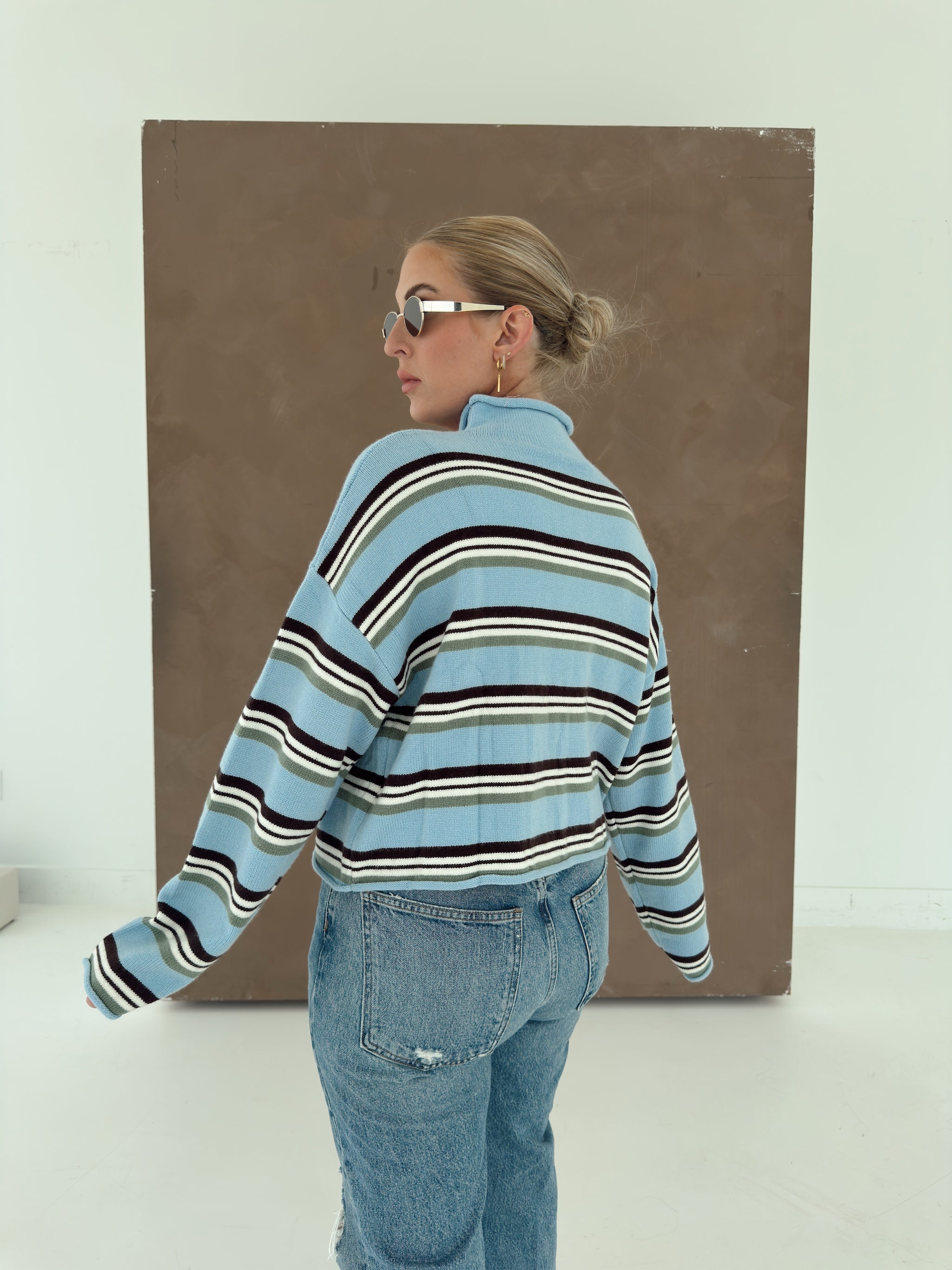 A woman wearing a light blue ribbed knit striped sweater with brown, white, and green stripes, paired with light-wash blue jeans, standing in front of a large brown canvas print.