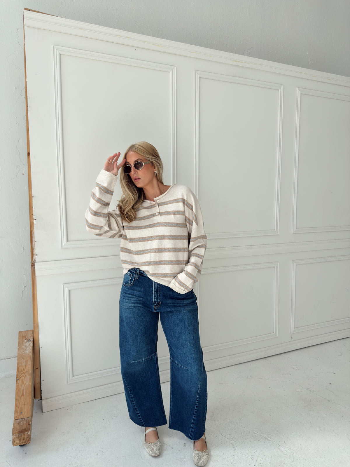 A woman stands in front of a white paneled wall wearing a beige, cream, and light blue horizontally striped long-sleeve knit top, wide-leg blue jeans, and light-colored flats.