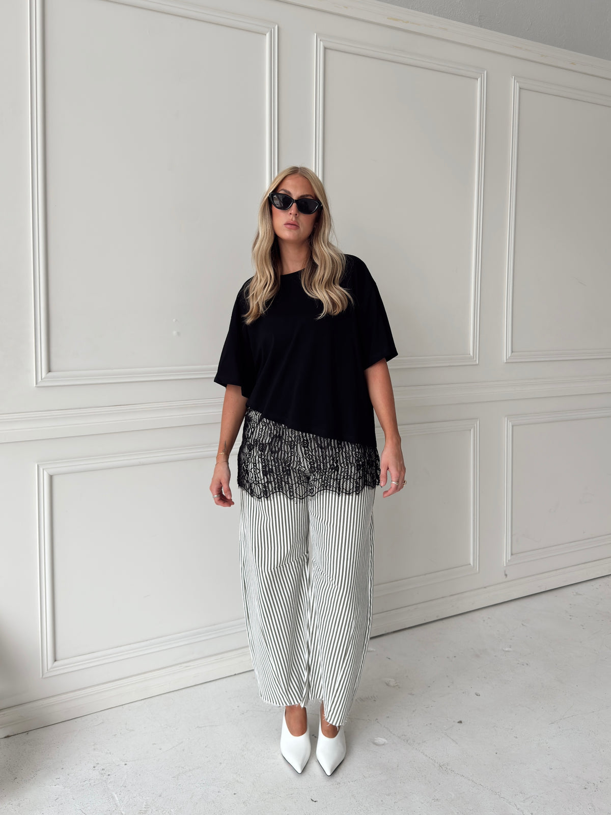 A woman modeling a black asymmetrical top with a sheer black lace hem, paired with white and olive vertically striped pants, standing in front of a white paneled studio backdrop.
