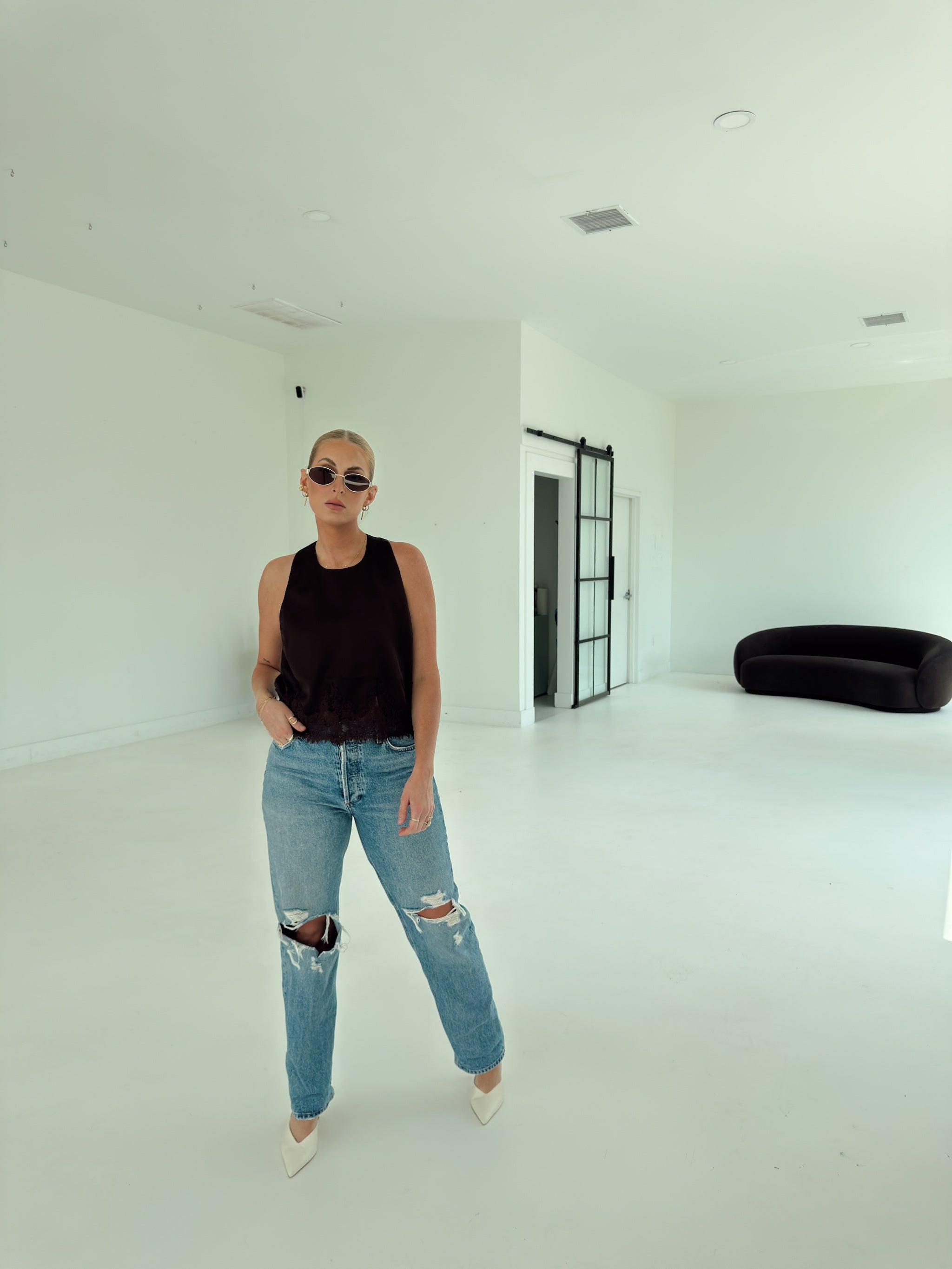 A woman wearing a brown sleeveless top, light blue ripped jeans, and white pointed shoes stands in a bright, minimalist white room with a black curved sofa in the background.