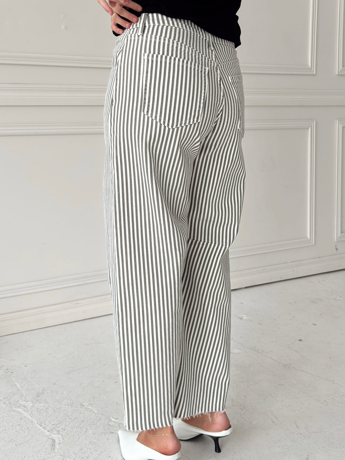 A woman modeling a pair of wide-leg barrel jeans with thin olive and white vertical stripes, paired with a black t-shirt, and white backless pointed-toe heels, standing in front of a white paneled wall.
