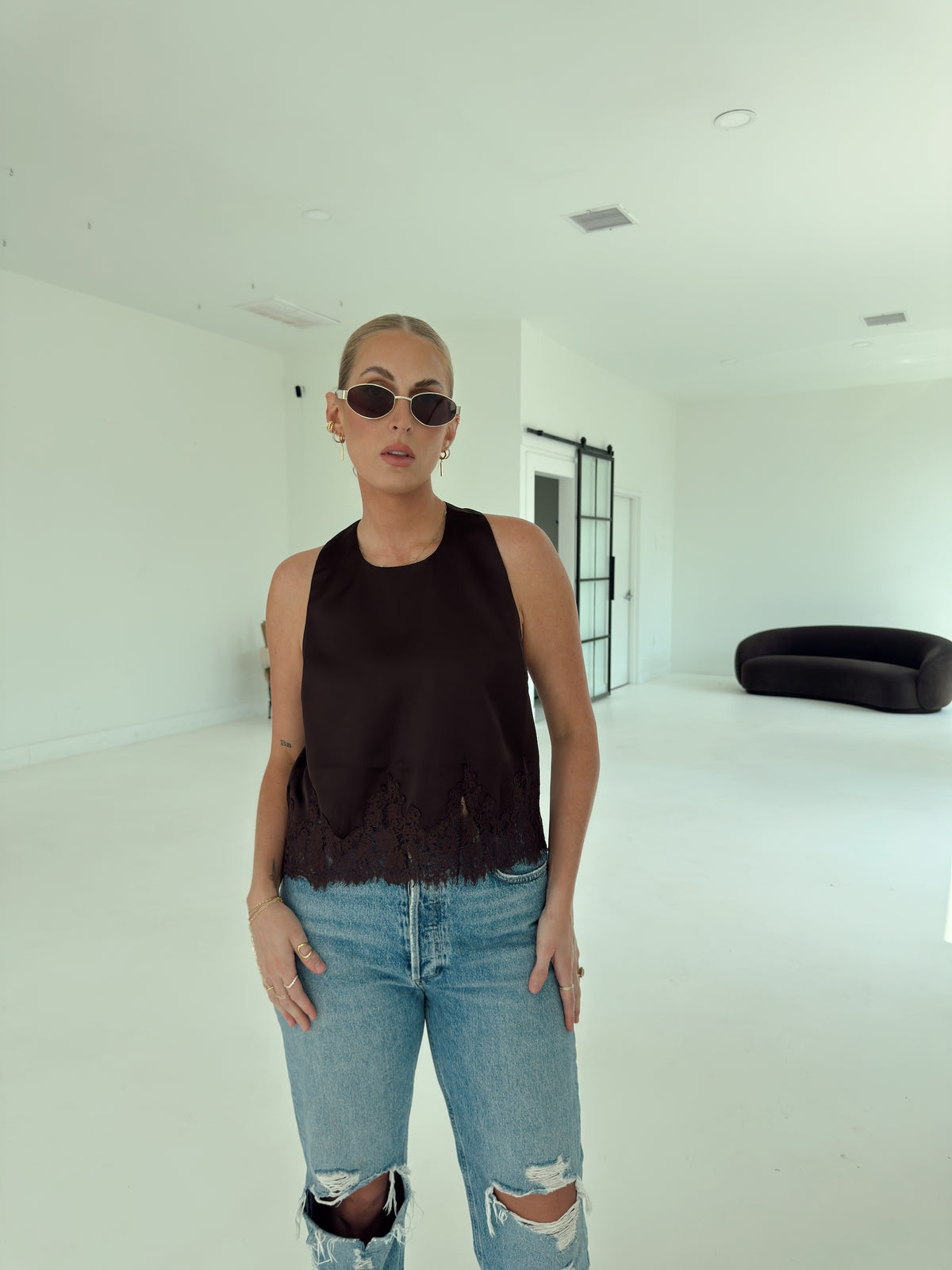 A woman wearing a brown sleeveless top with a frayed hem and light-wash distressed jeans, standing in a bright, minimalist white room.
