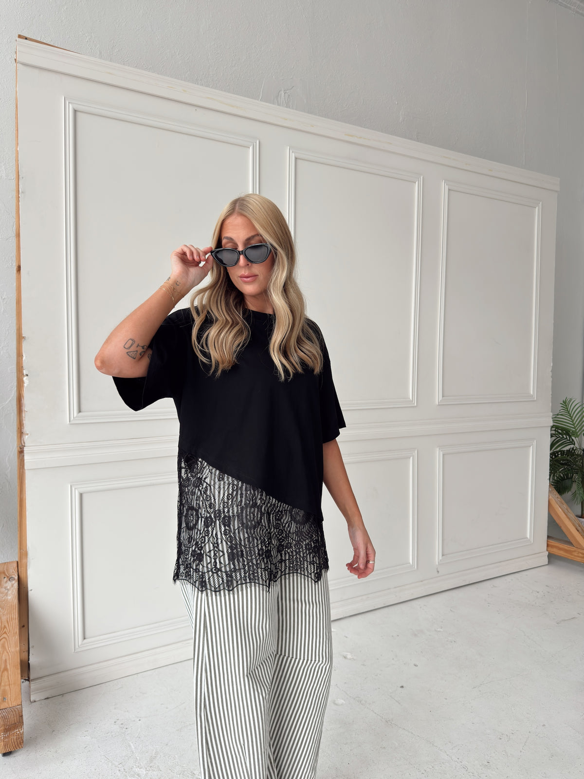 A woman modeling a black asymmetrical top with a sheer black lace hem, paired with white and olive vertically striped pants, standing in front of a white paneled studio backdrop.