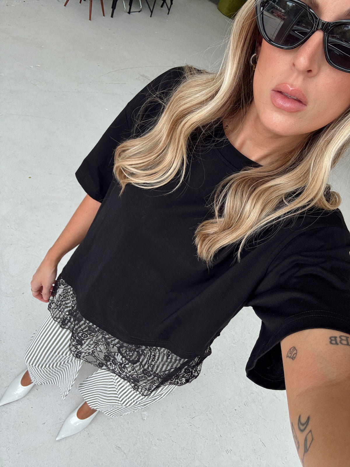 A woman wearing a black short-sleeve t-shirt with a sheer black lace hem, paired with olive and white striped pants and white pointed shoes, photographed in a casual indoor setting.
