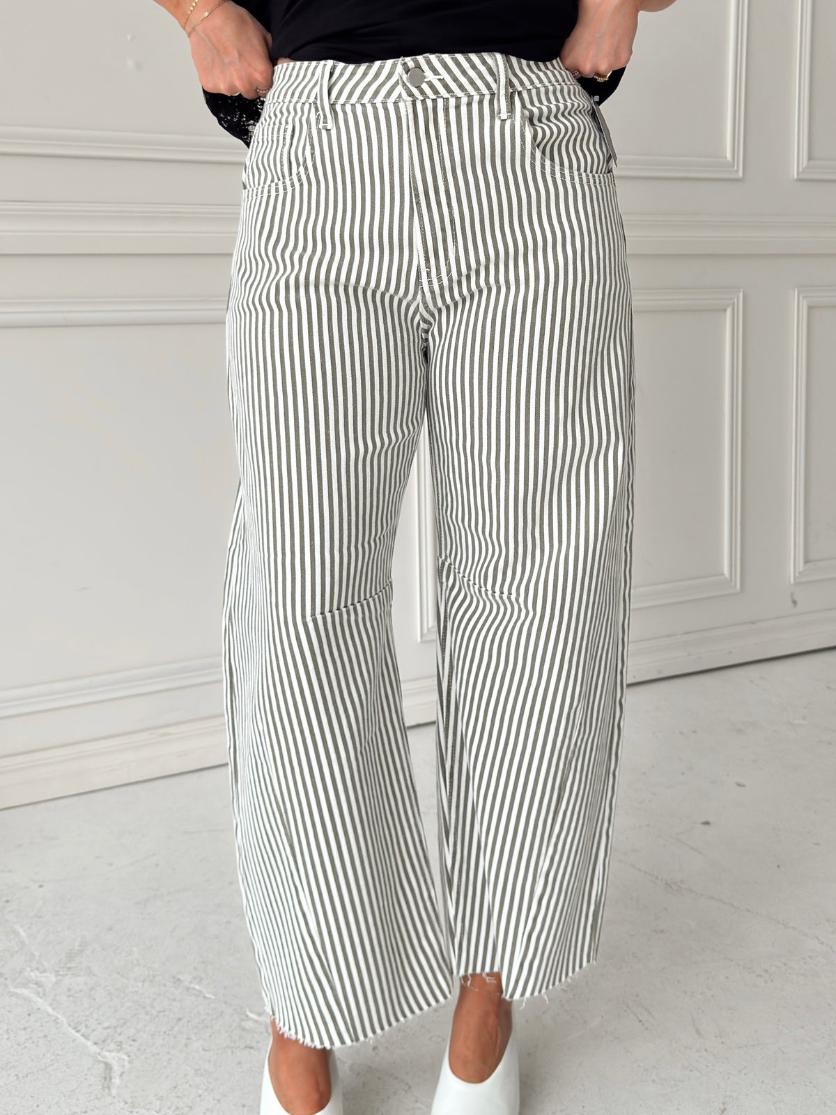 A woman modeling a pair of wide-leg barrel jeans with thin olive and white vertical stripes, paired with a black t-shirt, and white pointed-toe heels, standing against a white paneled wall.