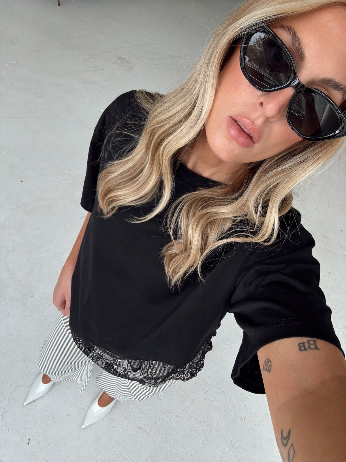 A woman wearing a black short-sleeve t-shirt, black with olive and white striped pants with a black lace hem, and white pointed-toe heels, photographed in a casual selfie-style overhead shot against a light gray concrete floor.