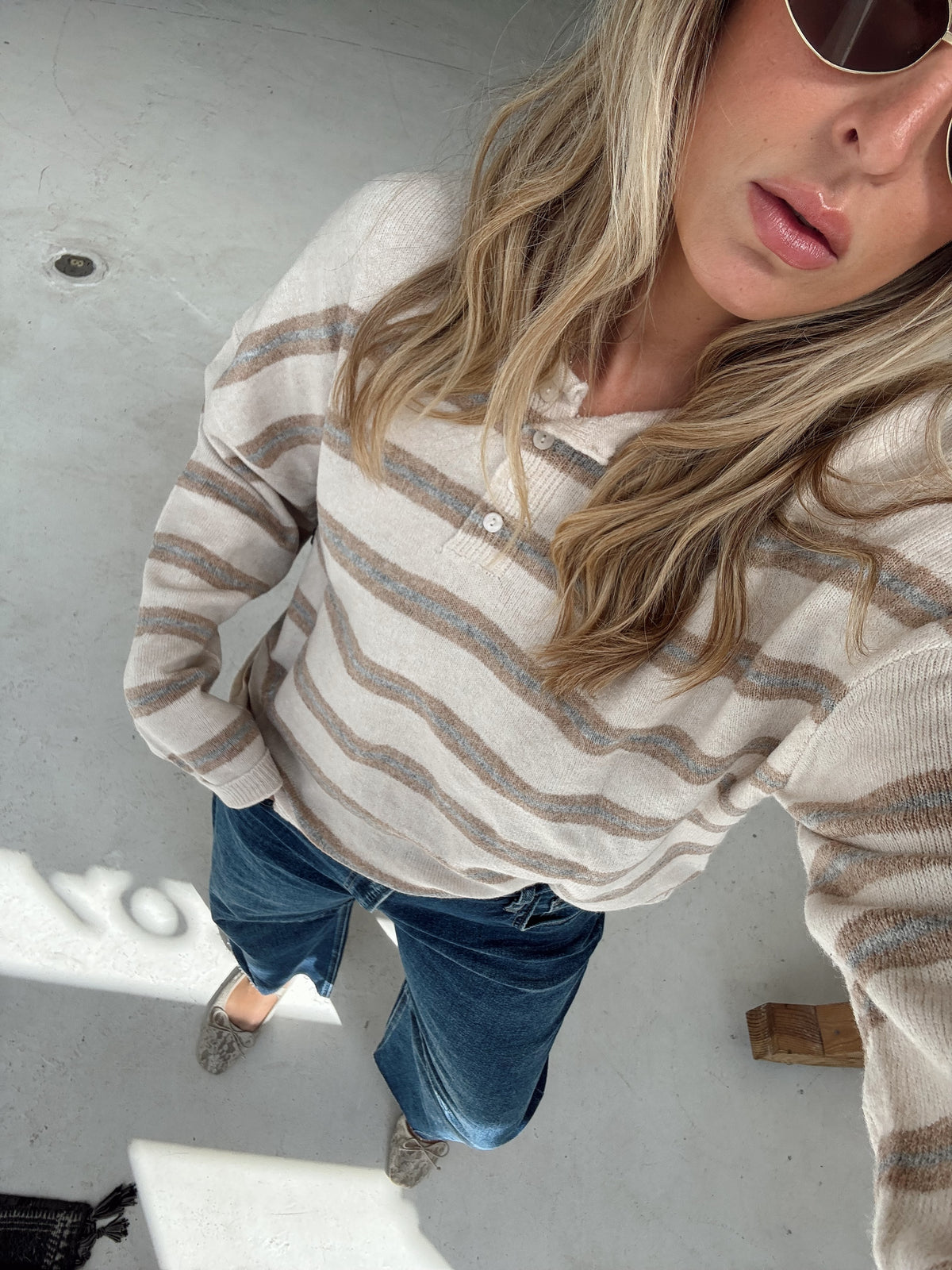 A woman wearing a beige, light blue, and cream striped long-sleeve knit sweater, blue jeans, and light slip-on shoes, photographed from a high-angle selfie perspective on a concrete floor.
