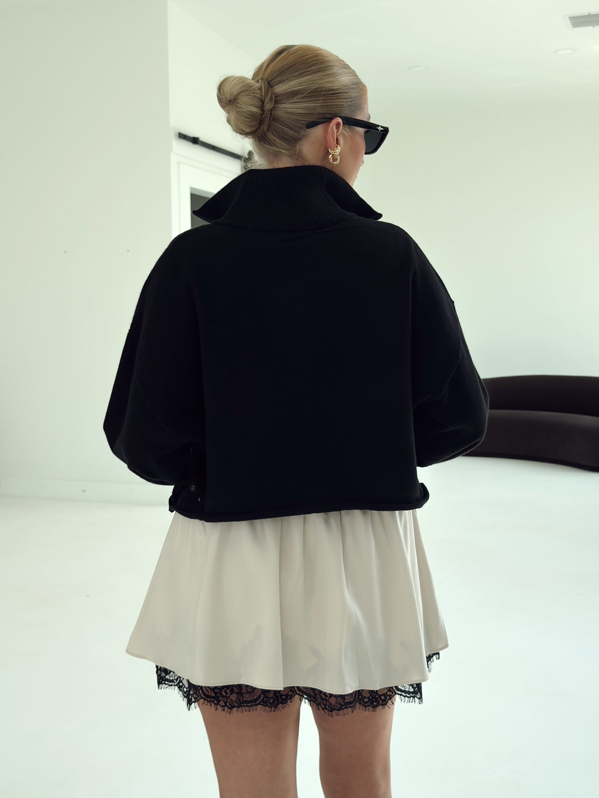 A woman viewed from behind wearing a black cropped long-sleeve jacket over an off-white mini skirt with a black lace hem, in a bright minimalist room.