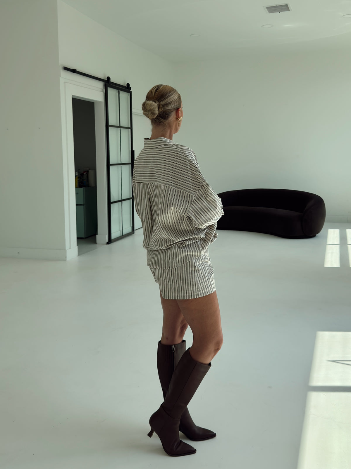 A woman stands in a minimalist white room wearing a brown and white vertically striped shirt and matching striped mini skirt, paired with dark brown knee-high heeled boots.