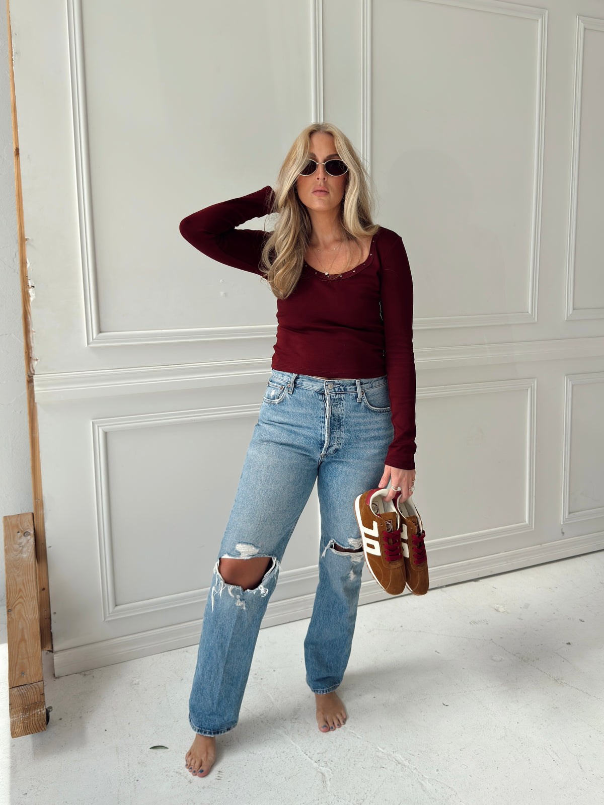 A woman stands barefoot in a white paneled room wearing a burgundy long-sleeve top, light-wash distressed blue jeans, and holding a pair of brown and white sneakers.