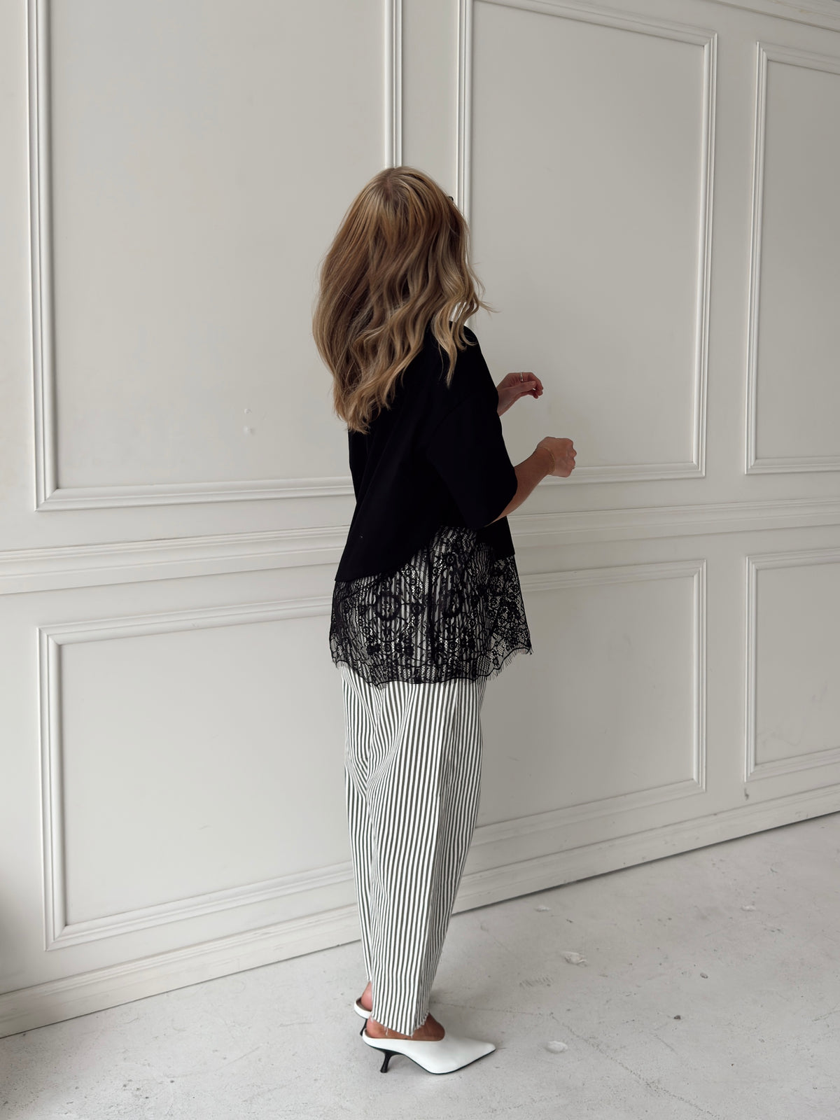 A woman stands with her back to the camera wearing a black top with a sheer black lace overlay, white and olive vertically striped jeans, and white pointed-toe mule heels, set against a white paneled wall.