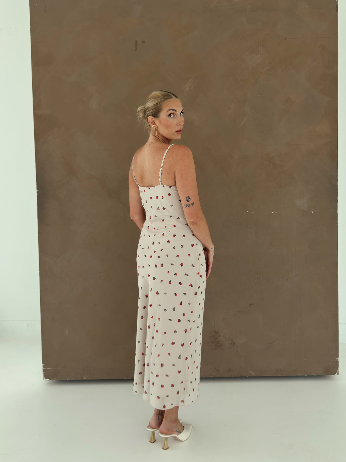 A woman stands with her back to the camera wearing a  beige colored maxi dress with a red and green floral pattern, paired with white mule heels, set against a textured brown backdrop.