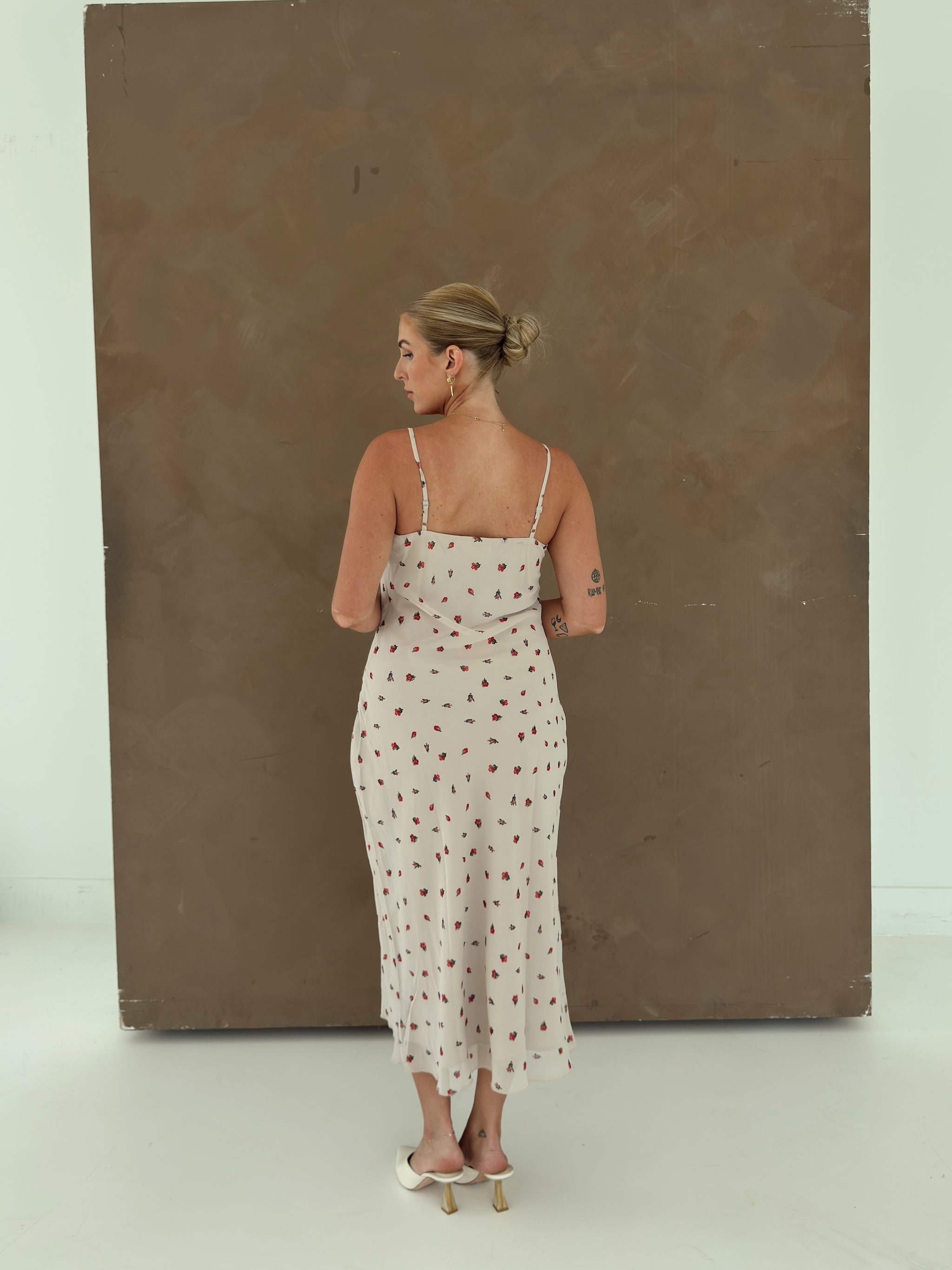 A model with her back to camera wearing a beige spaghetti-strap slip dress with small red ditsy floral print and lace trim.
