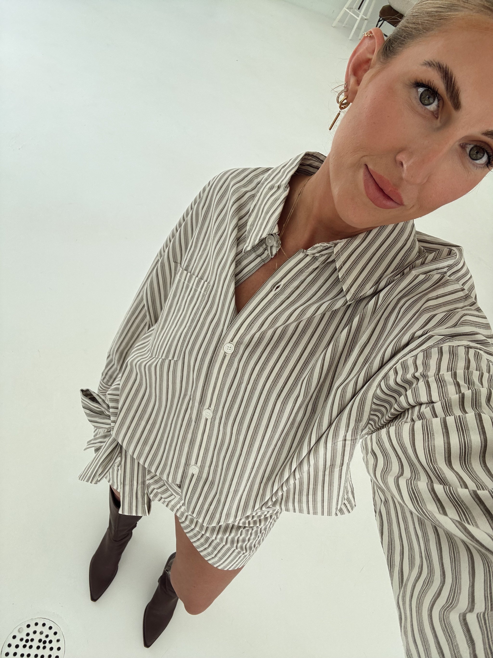 A woman wearing an oversized long-sleeve button-up shirt with vertical brown and white stripes, paired with dark brown pointed-toe ankle boots, photographed in a high-angle selfie on a white floor.