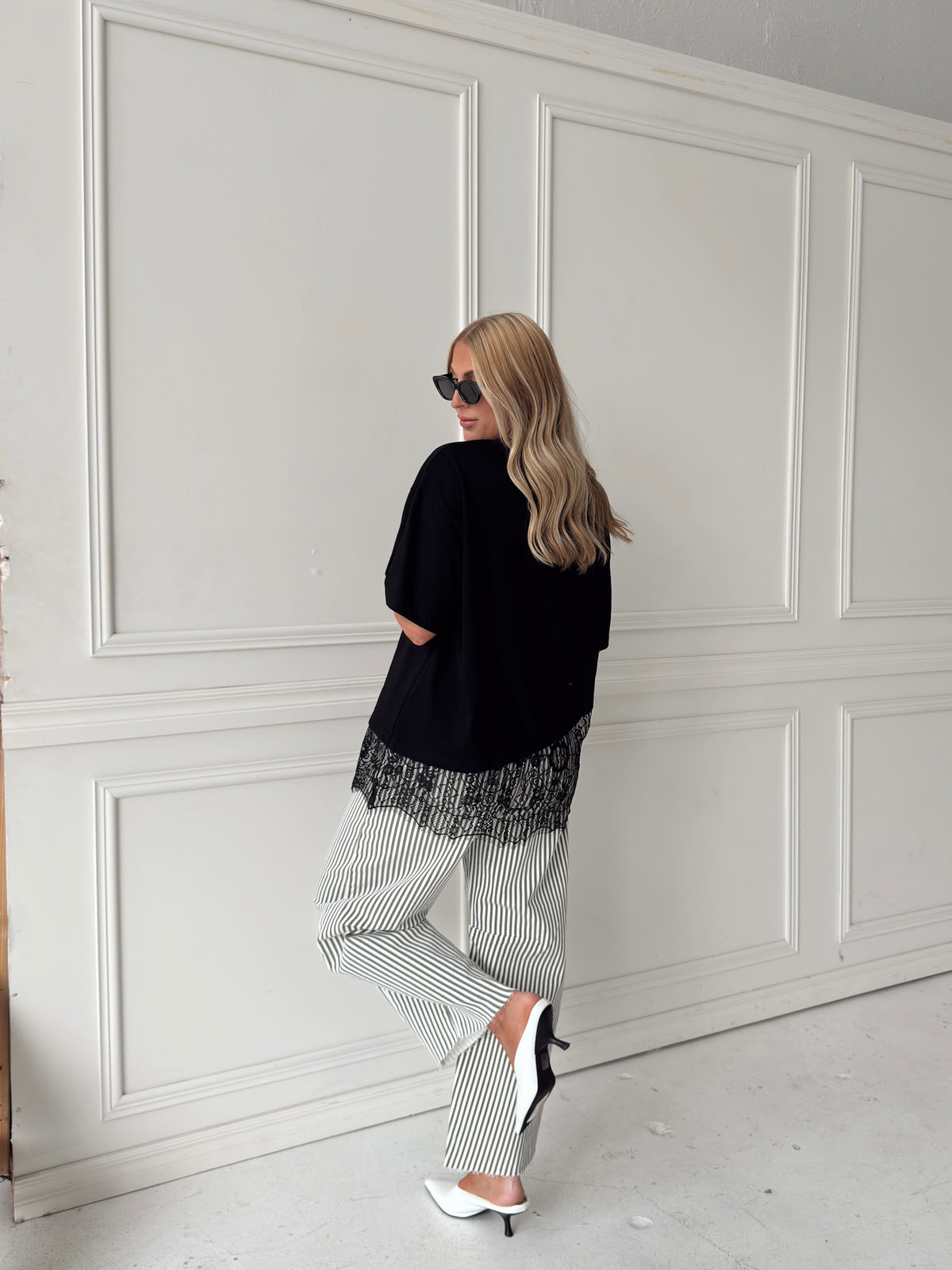 A woman modeling a black asymmetrical top with a sheer black lace hem, paired with white and olive vertically striped pants, standing in front of a white paneled studio backdrop.