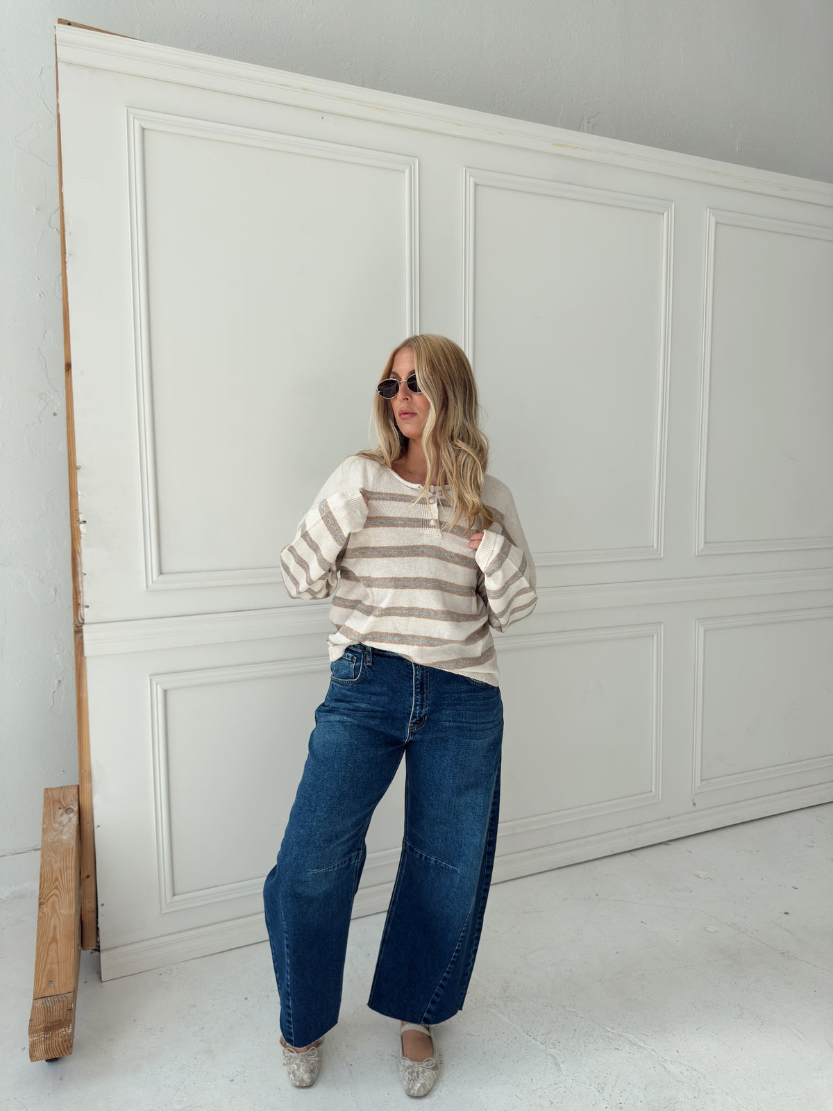 A woman stands in front of a white paneled wall wearing a beige, cream, and light blue horizontally striped long-sleeve knit top, wide-leg blue jeans, and light-colored flats.