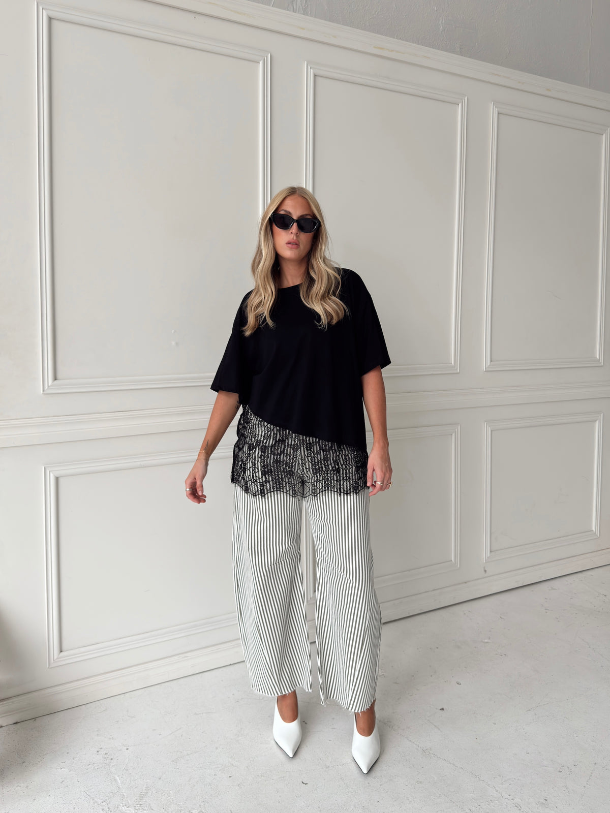 A woman modeling a black asymmetrical top with a sheer black lace hem, paired with white and olive vertically striped pants, standing in front of a white paneled studio backdrop.