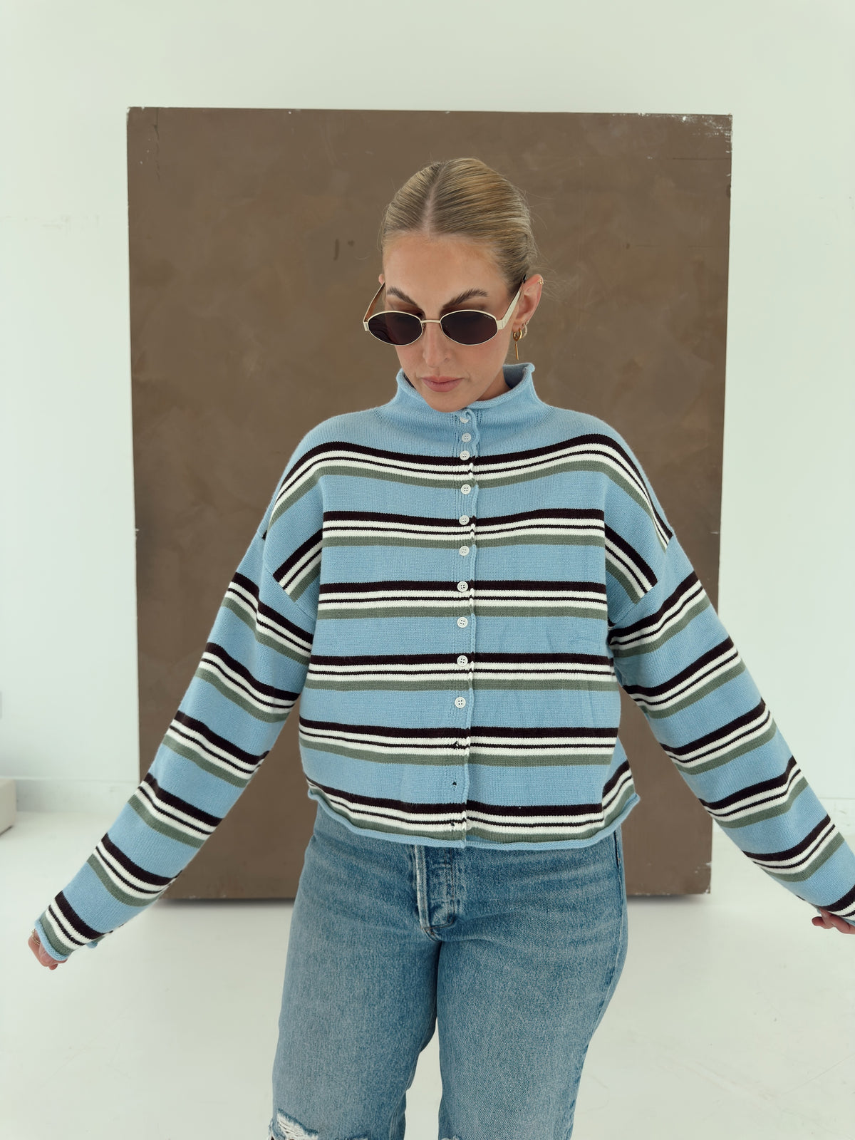A person wearing a light blue knit cardigan with horizontal brown, white, and olive green stripes, paired with light-wash jean.