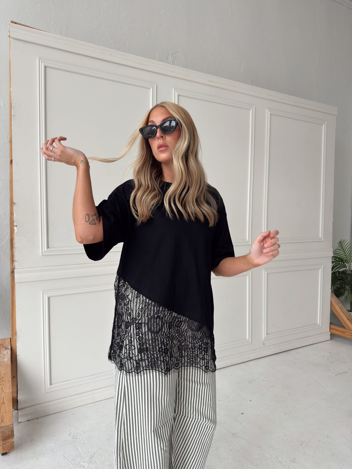 A woman modeling a black asymmetrical top with a sheer black lace hem and striped pants, posing in front of a white paneled wall.