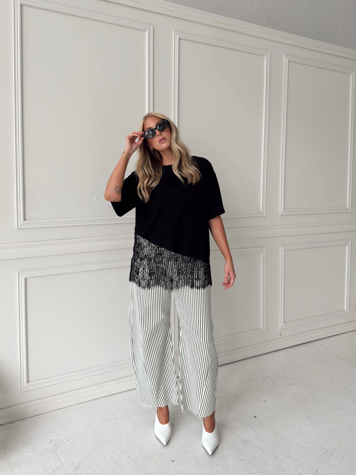 A woman modeling a black asymmetrical top with a sheer black lace hem, paired with white and olive vertically striped pants, standing in front of a white paneled studio backdrop.