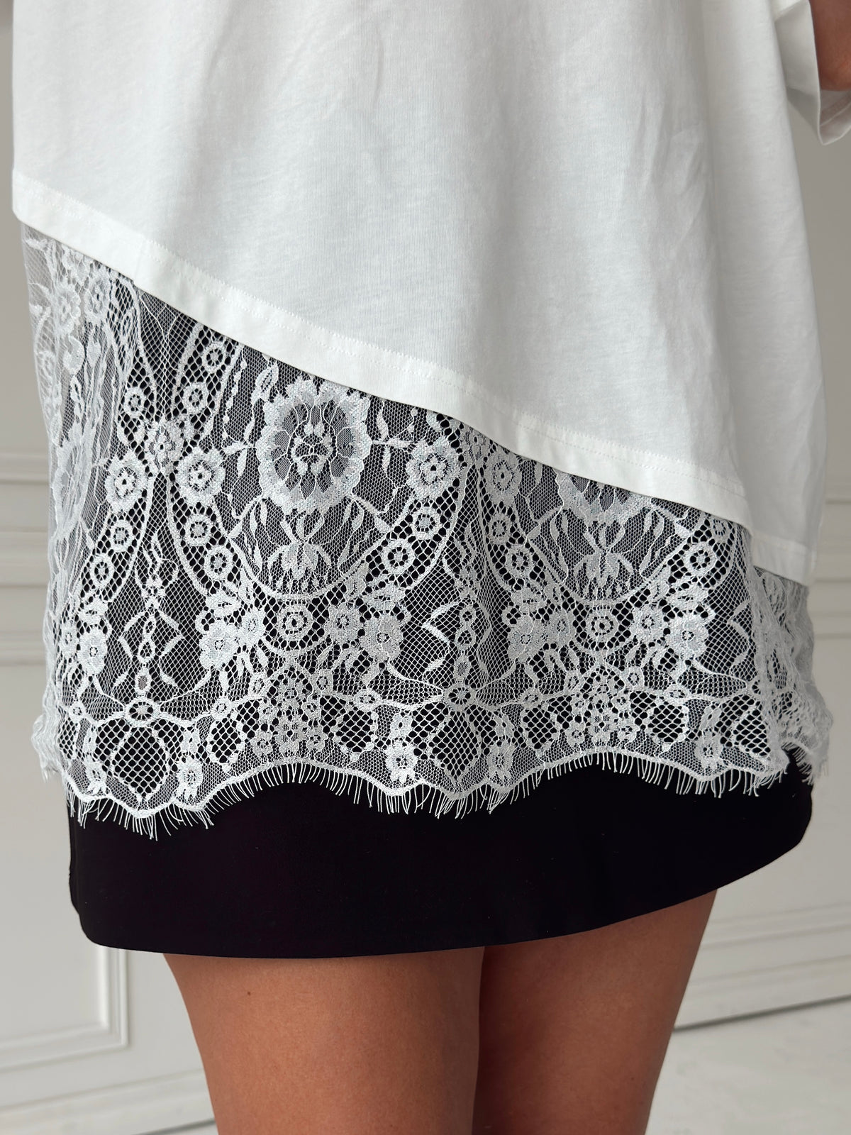 Close-up of the lower half of a person wearing a white top with a sheer white floral lace panel and a black skort or shorts underneath, standing indoors against a light paneled wall.