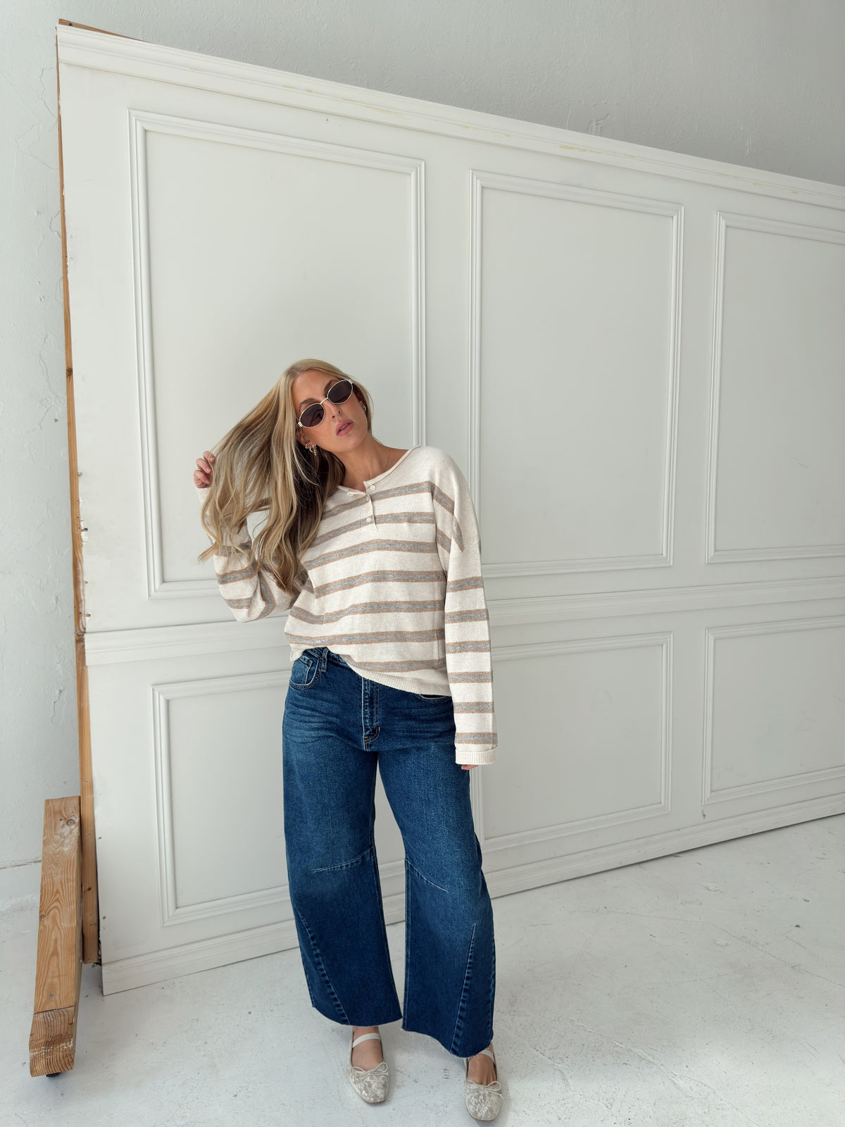 A woman stands in front of a white paneled wall wearing a beige, cream, and light blue horizontally striped long-sleeve knit top, wide-leg blue jeans, and light-colored flats.