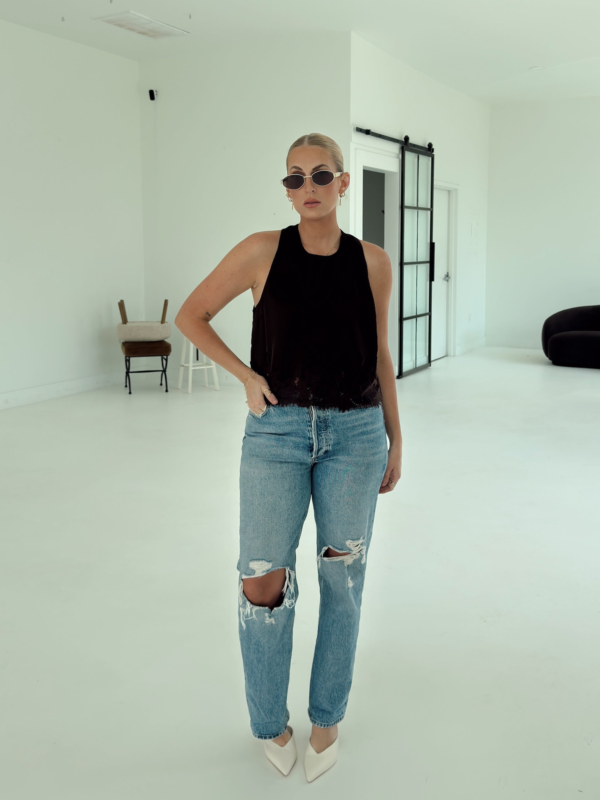 A woman stands in a bright white studio wearing a brown sleeveless top, light-wash distressed blue jeans with ripped knees, and white pointed heels.