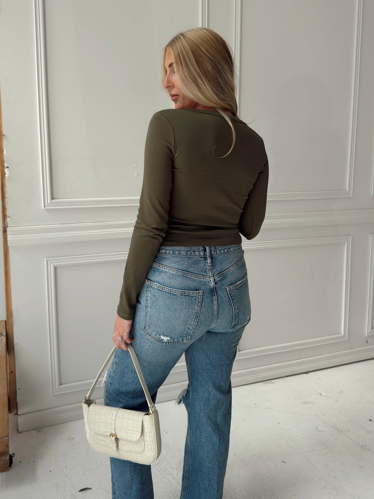 A woman is shown from behind wearing a fitted olive green long-sleeve top and light blue jeans, holding a small white handbag in a room with white paneled walls.