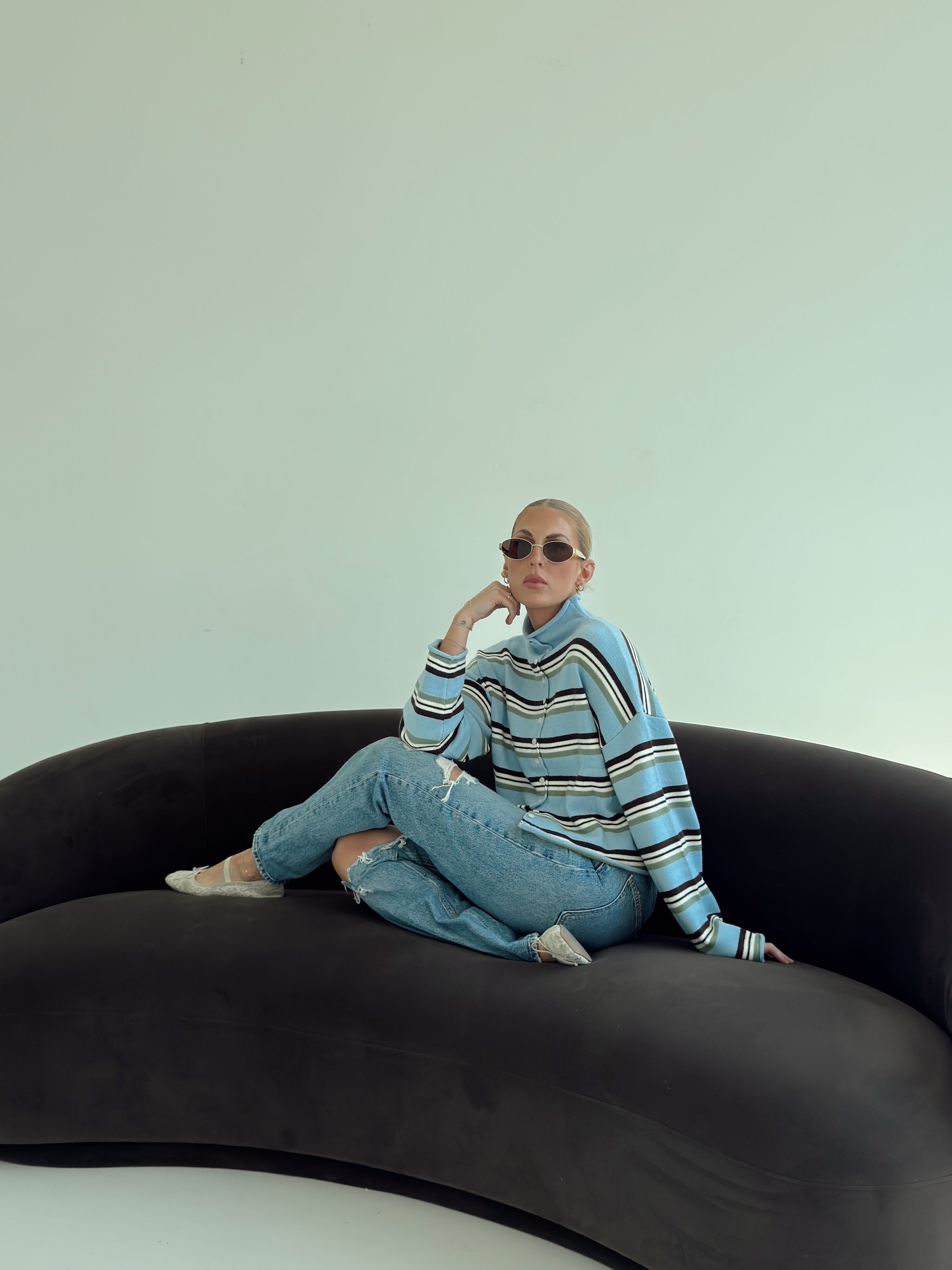 A model sits cross-legged on a dark curved sofa against a pale mint wall, wearing a light blue striped sweater, light-wash jeans, and white flats.