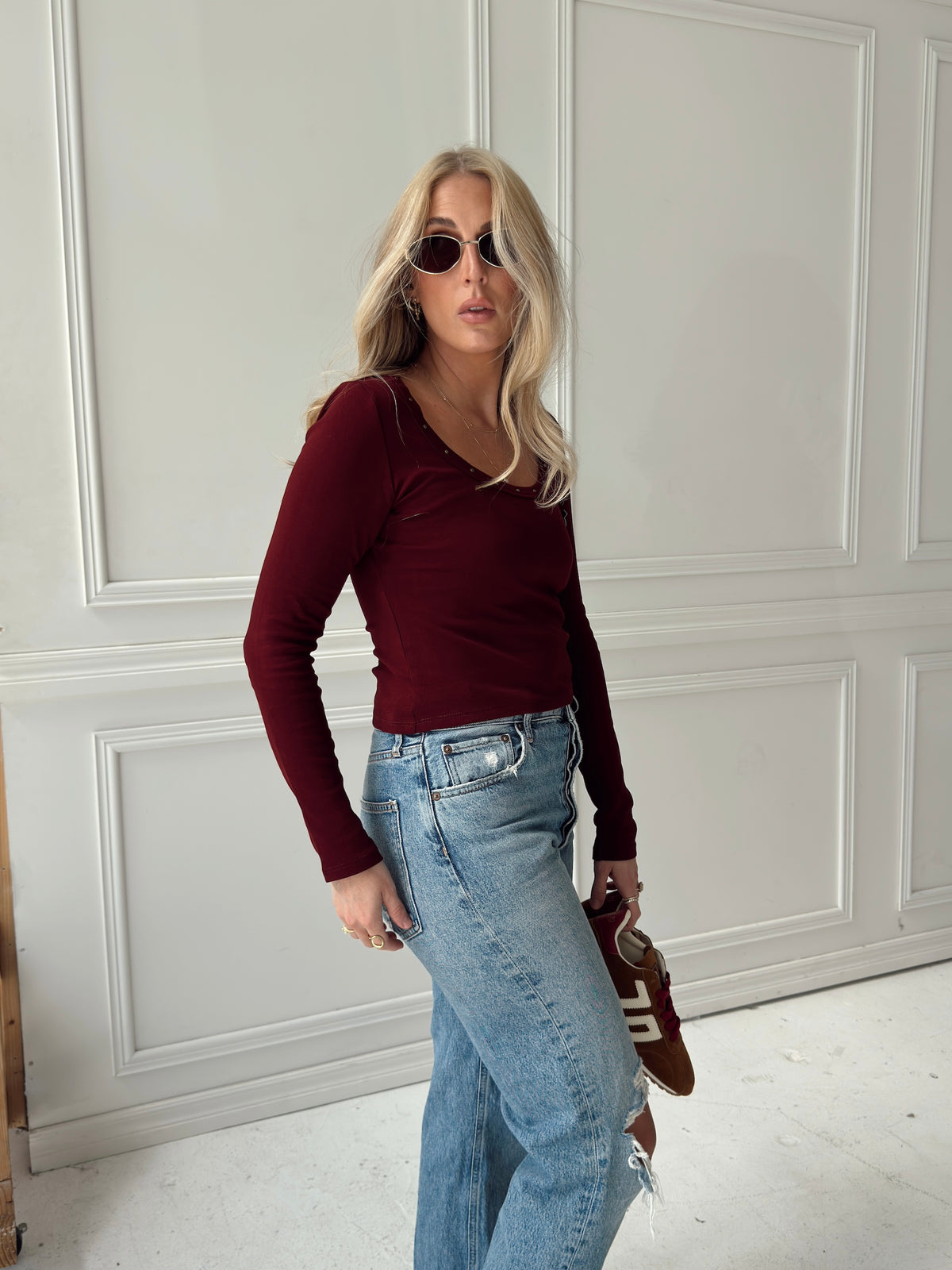 A woman with blonde hair wearing a long-sleeve maroon scoop-neck top, light-wash blue jeans, and sunglasses, holding a brown leather shoes, standing in front of a white paneled wall.