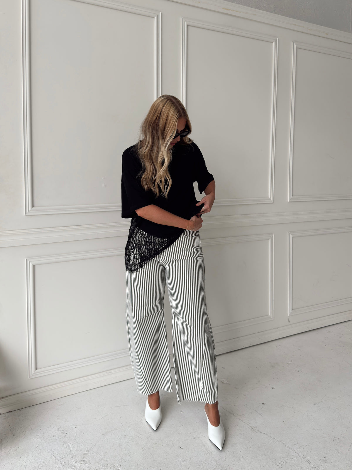 A woman modeling a pair of wide-leg barrel jeans with thin olive and white vertical stripes, paired with a black t-shirt, black sunglasses, and white pointed-toe heels, standing in front of a white paneled wall.