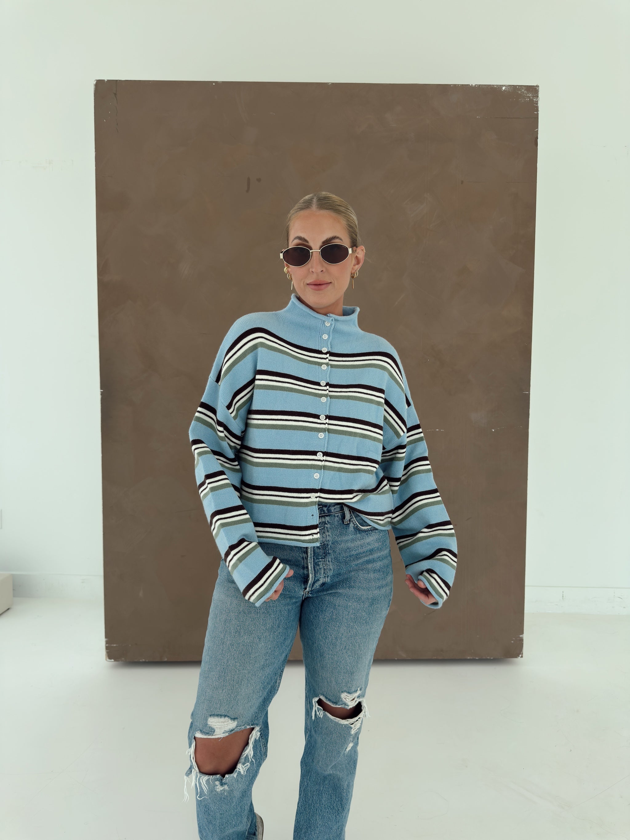 A woman wearing a light blue striped mock-neck knit sweater and ripped blue jeans, standing in front of a large brown rectangular art piece on a white studio floor.
