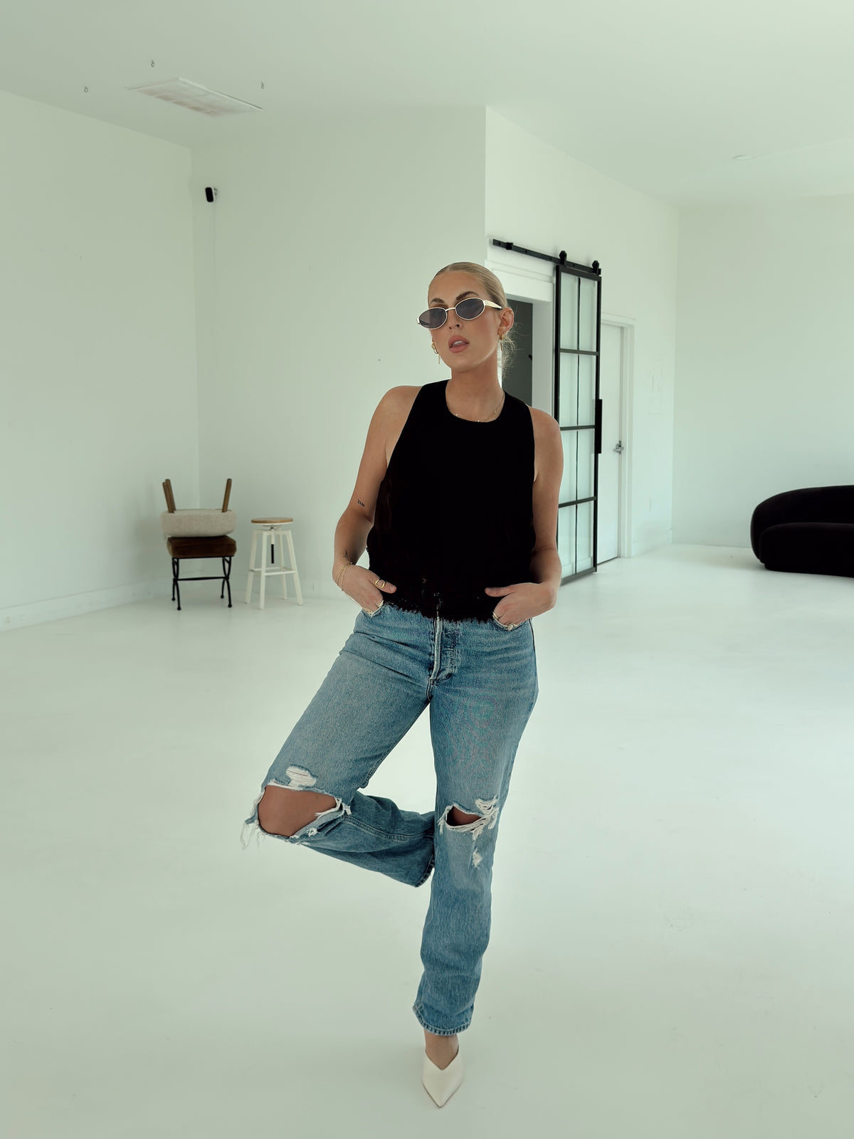 A woman posing in a minimalist white studio space, wearing a brown sleeveless top, light-wash distressed blue jeans, and white pointed-toe heels.