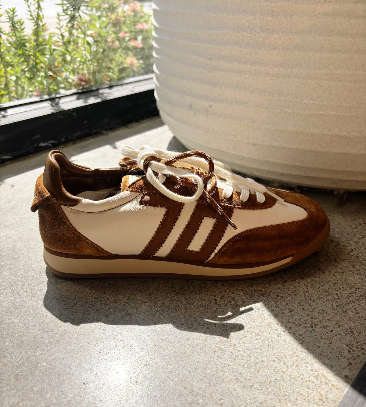 A low-top sneaker with brown suede and off-white leather panels, featuring three bold side stripes and cream-colored laces, resting on a speckled floor next to a large white ribbed planter.