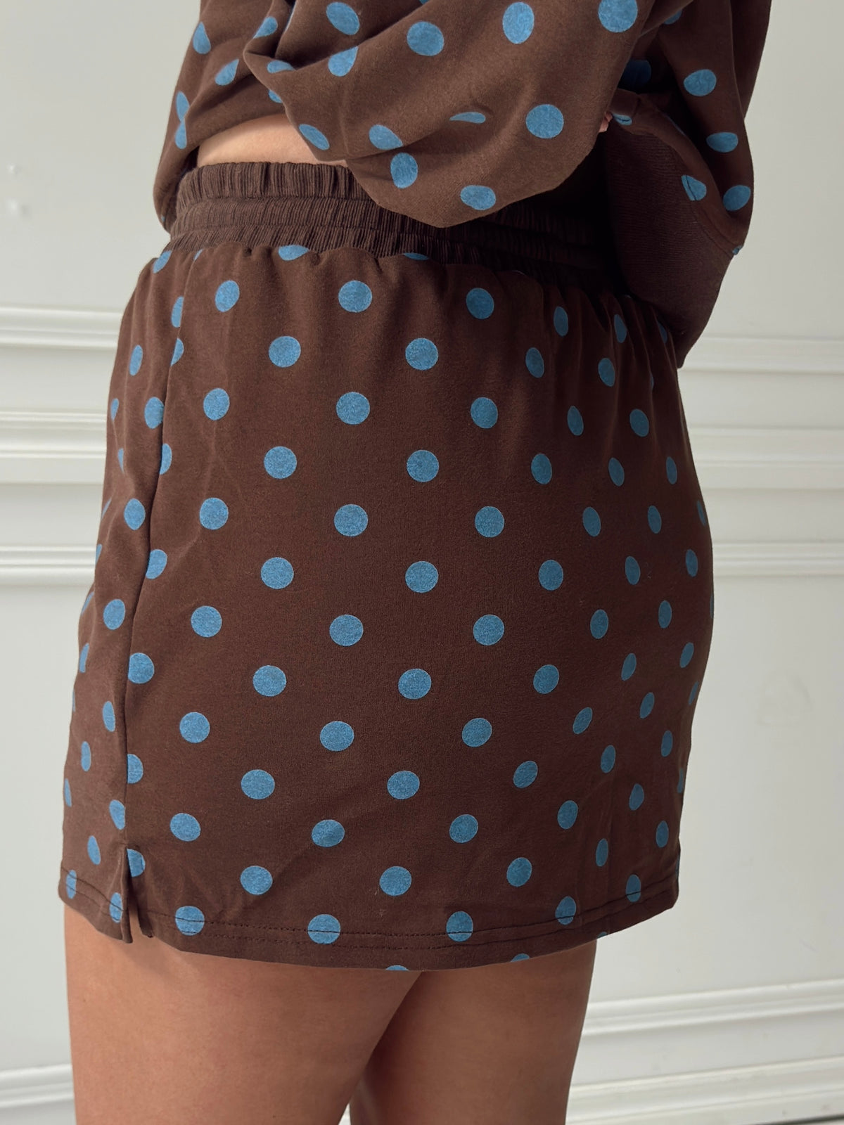 A close-up of a person wearing a brown pajama or lounge skort with light blue polka dots, paired with a matching top, set against a white paneled wall.
