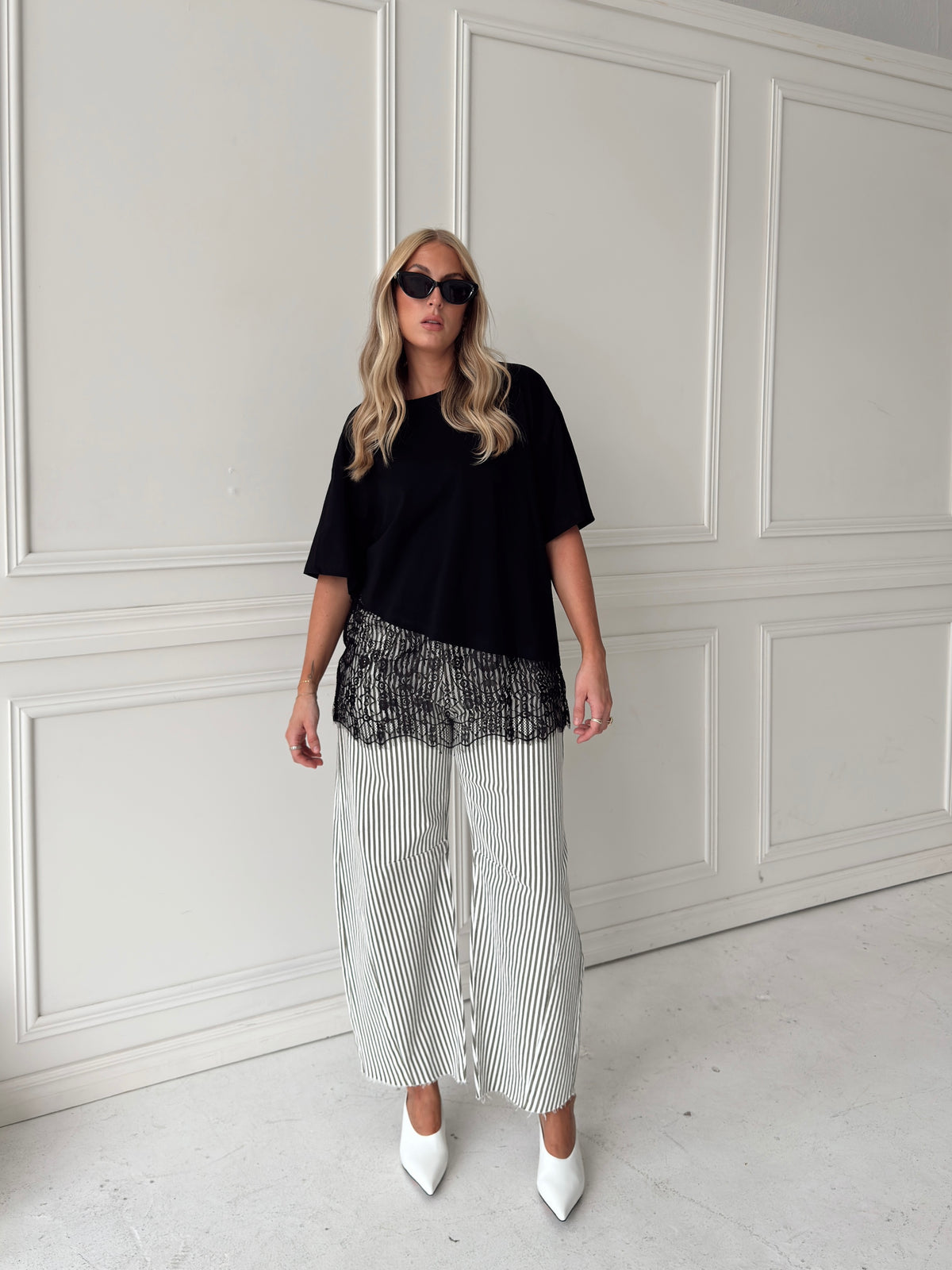A woman modeling a black asymmetrical top with a sheer black lace hem, paired with white and olive vertically striped pants, standing in front of a white paneled studio backdrop.