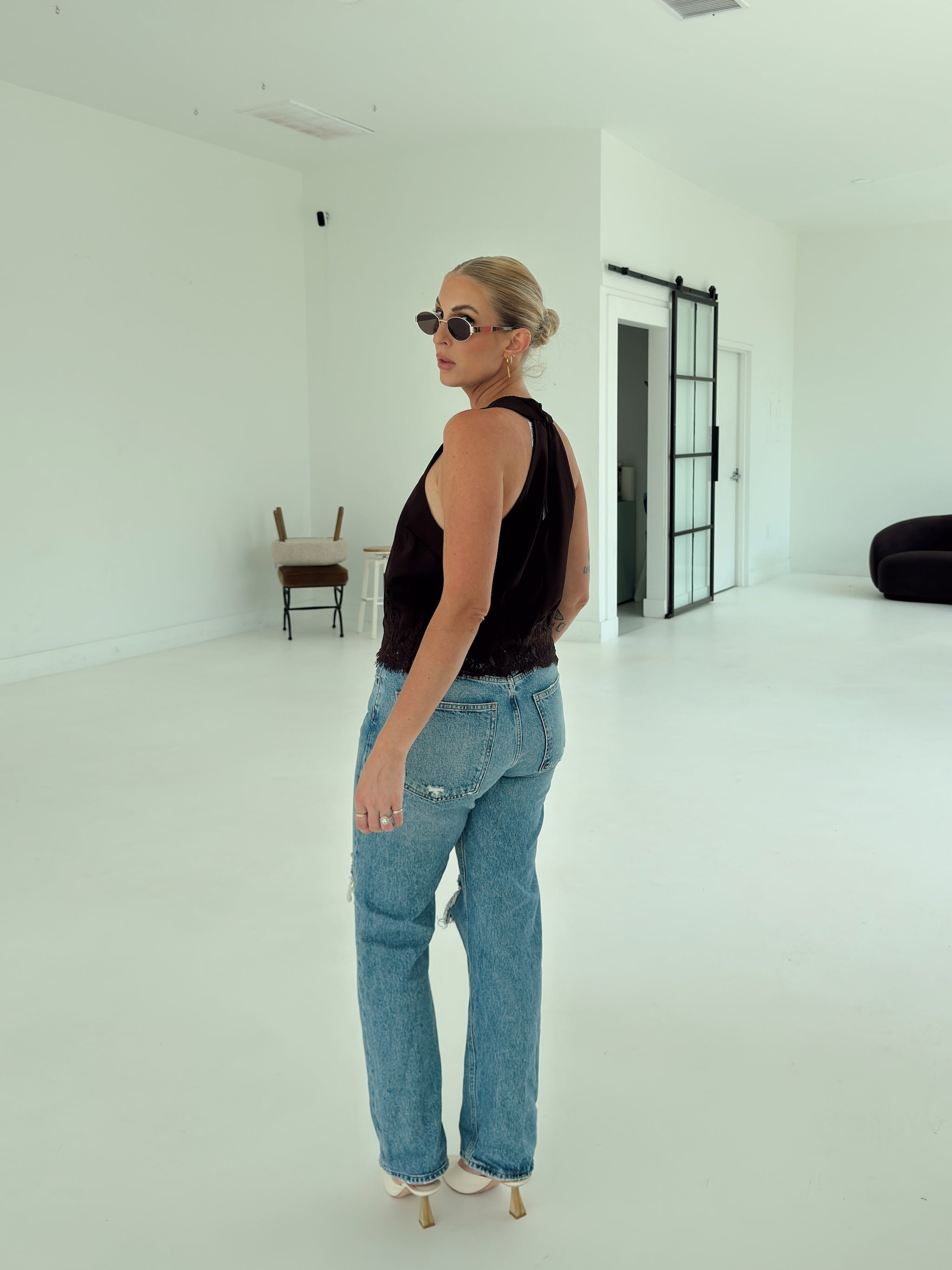 A woman wearing a brown sleeveless top and light blue straight-leg jeans stands in a bright, minimalist white studio space, looking over her shoulder.