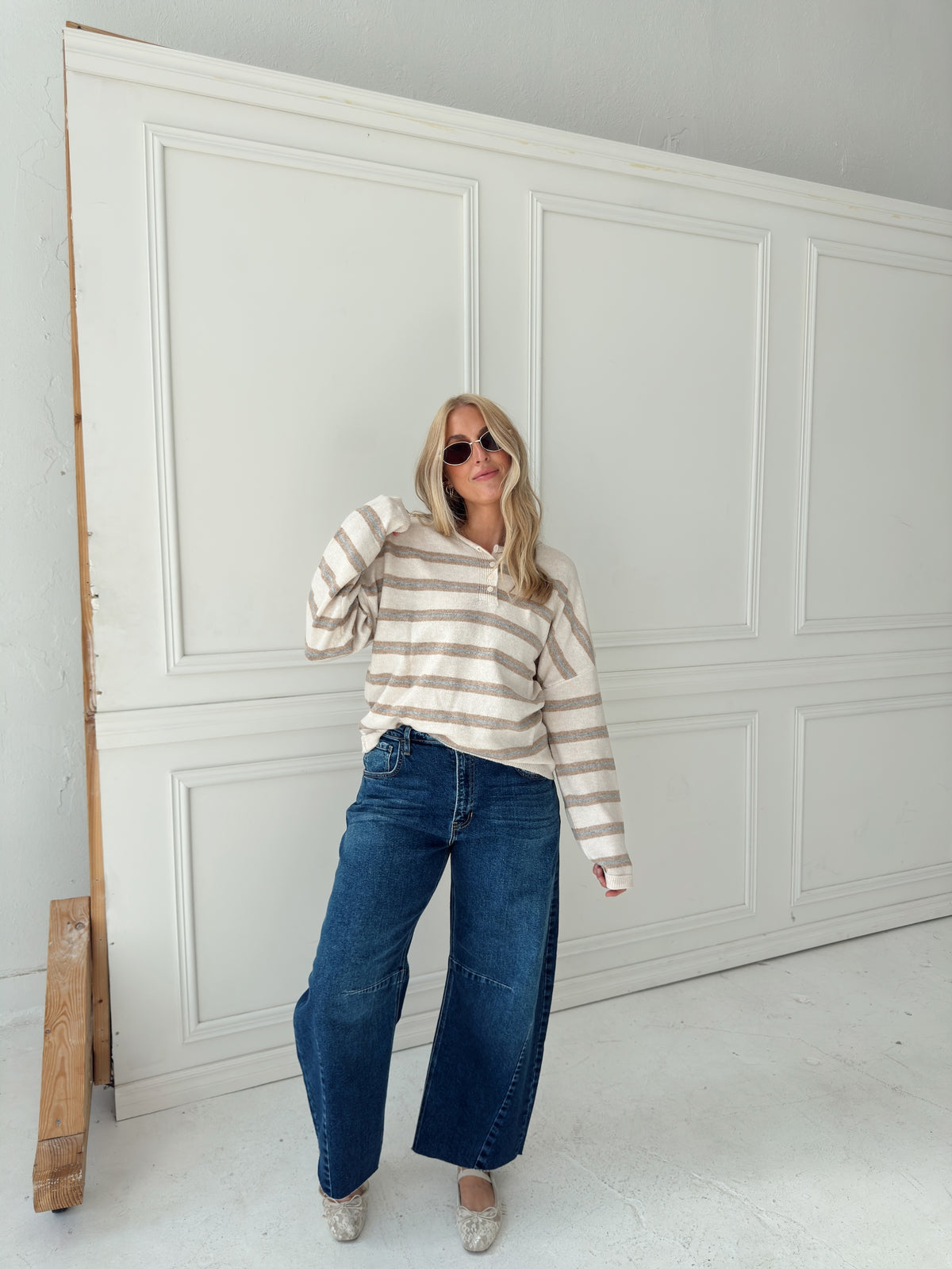 A woman wearing an oatmeal, cream, and light blue striped long-sleeve top, dark blue wide-leg jeans, and beige flats poses in front of a white paneled wall.