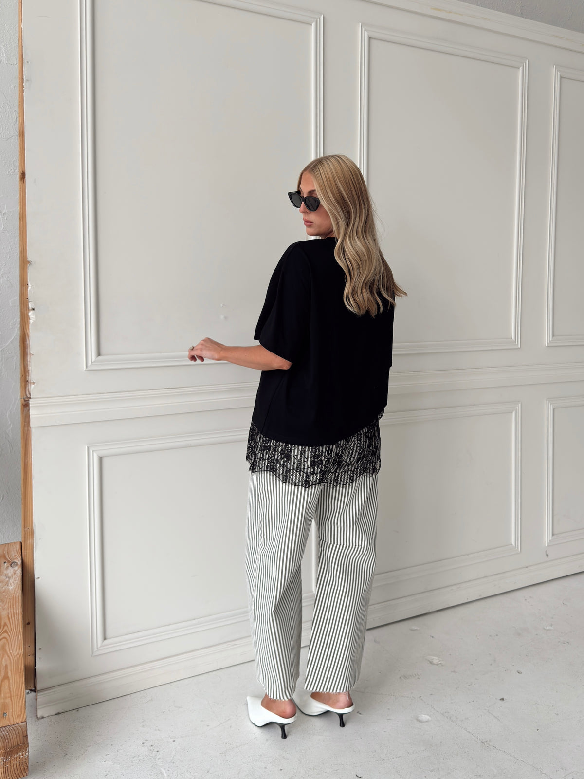 A woman modeling a pair of wide-leg barrel jeans with thin olive and white vertical stripes, paired with a black t-shirt, black sunglasses, and white pointed-toe heels, standing against a white paneled wall.