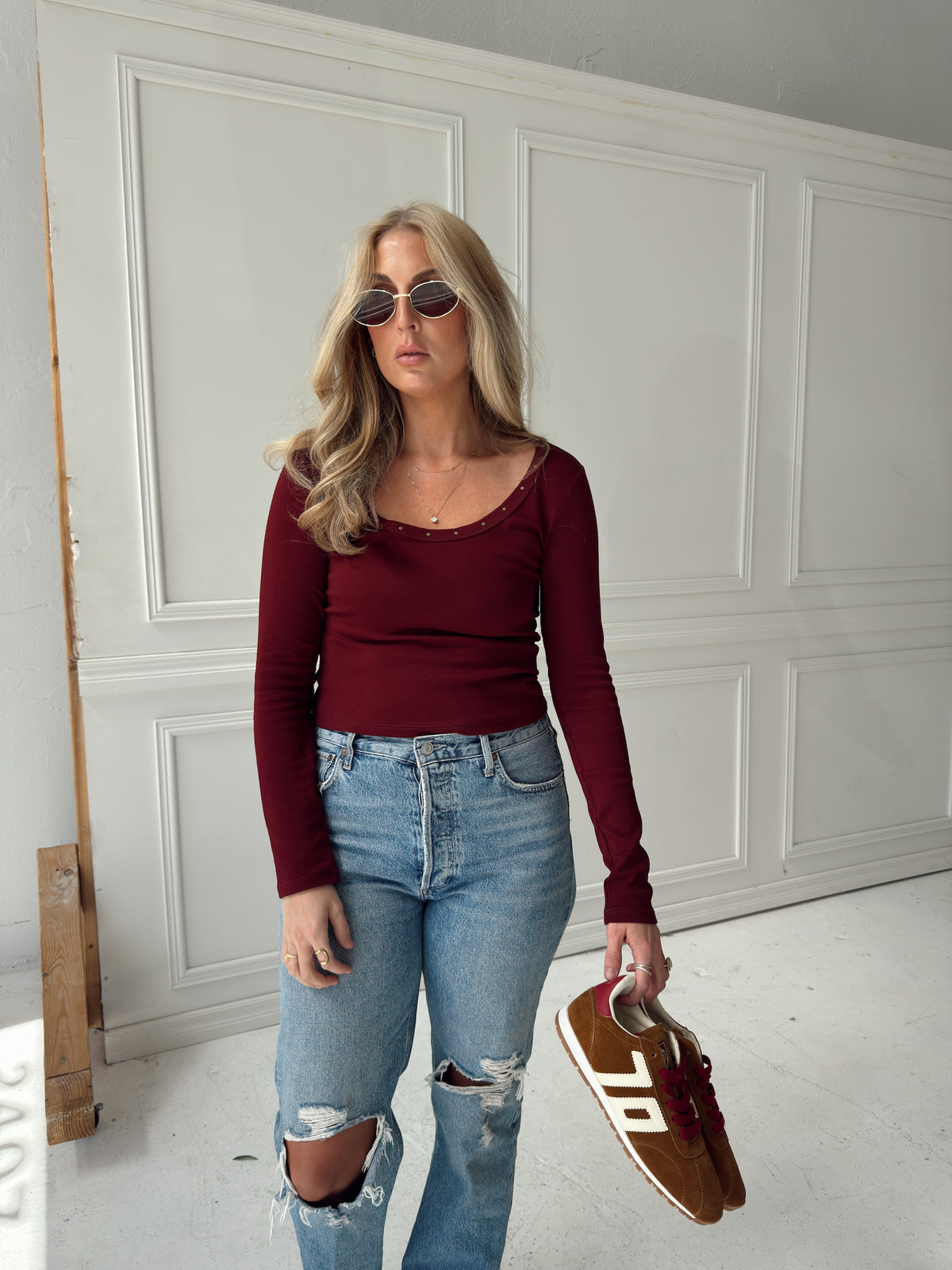 A woman modeling a long-sleeve burgundy scoop-neck top, distressed light-wash blue jeans, and sunglasses, holding brown suede sneakers in a bright studio with a white paneled wall background.