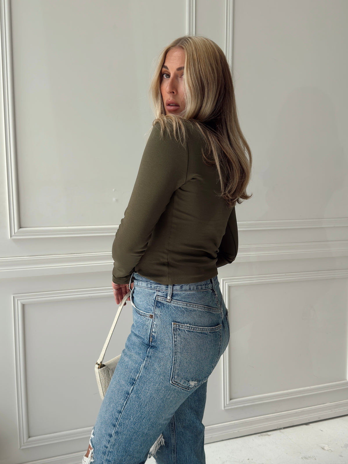 A woman with blonde hair is shown from a side-back angle, wearing an olive green long-sleeve top and light-wash blue jeans, holding a small white handbag against a white paneled wall.